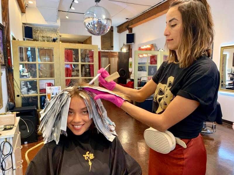 Woman with pink gloves styling a young girl's hair with foil rollers in a salon, smiling, interior with mirrors and decor.