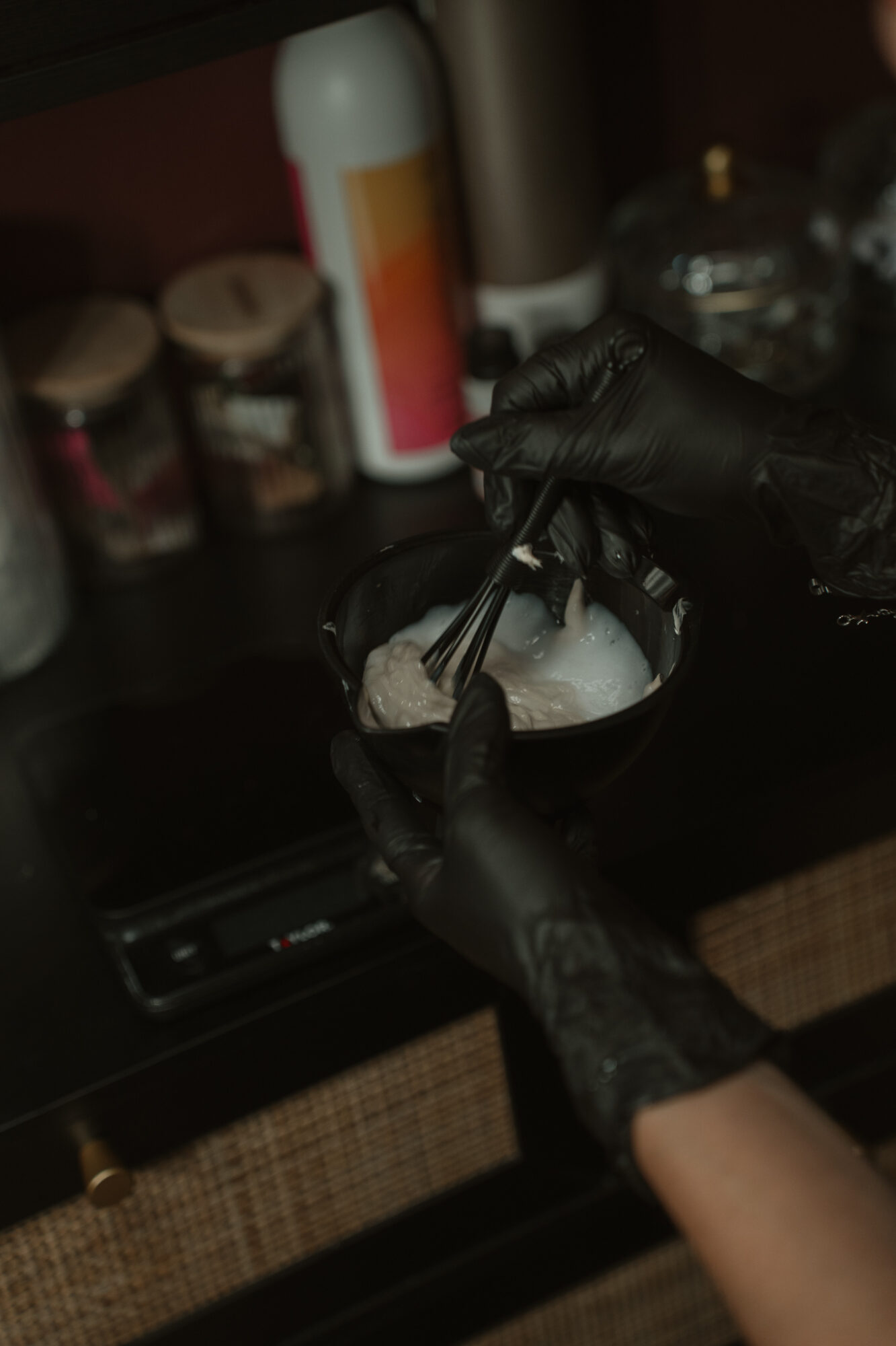 Person wearing black gloves mixing ingredients in a bowl with a spoon, in a kitchen setting with jars and bottles in background.