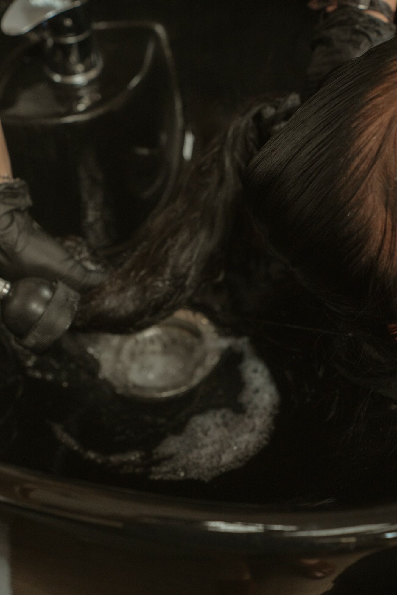 Person's hair being washed in a black sink with soap suds, viewed from above, with dark surroundings.