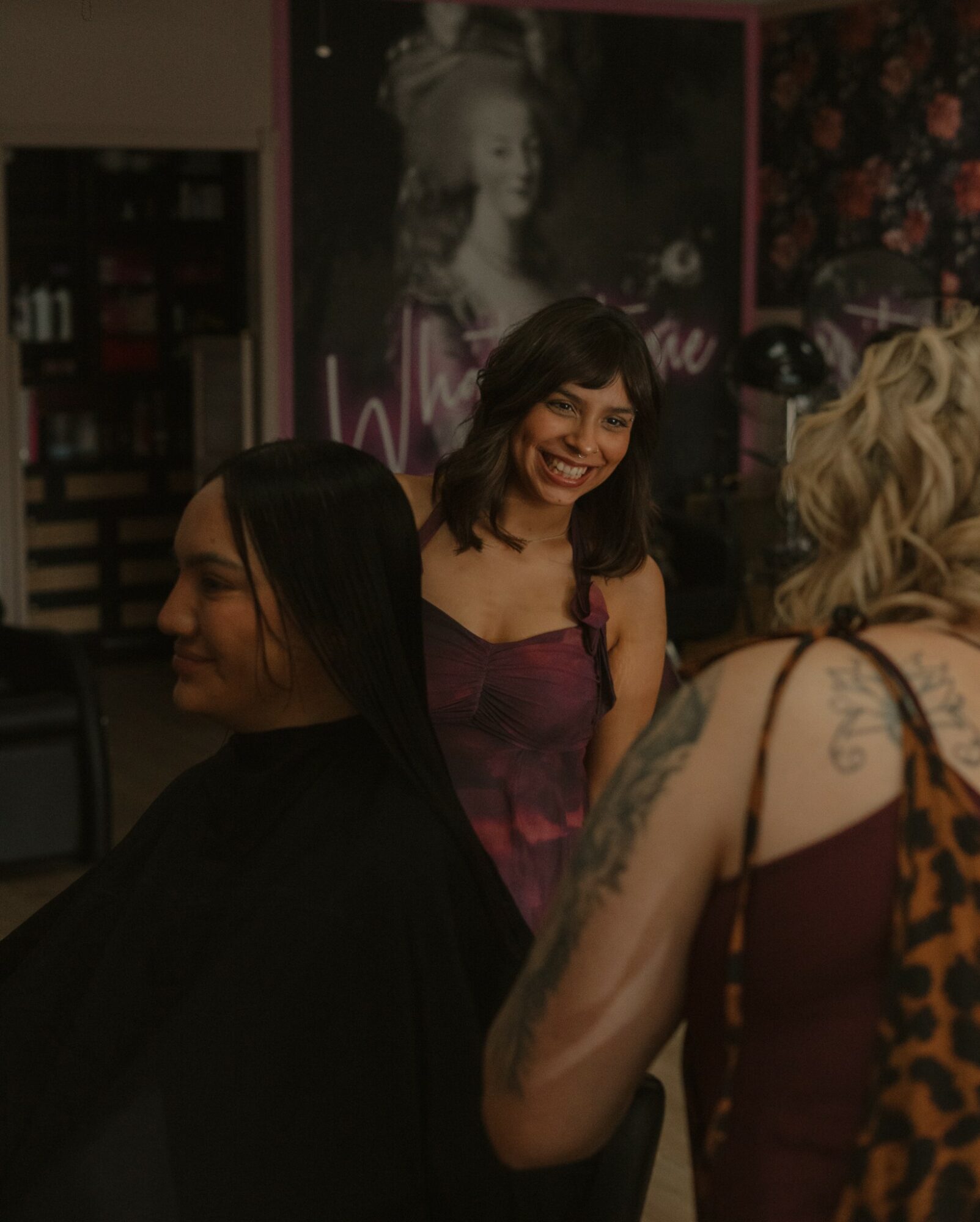 Three women talking in a dimly lit room, one smiling, with a poster and floral wallpaper in the background.