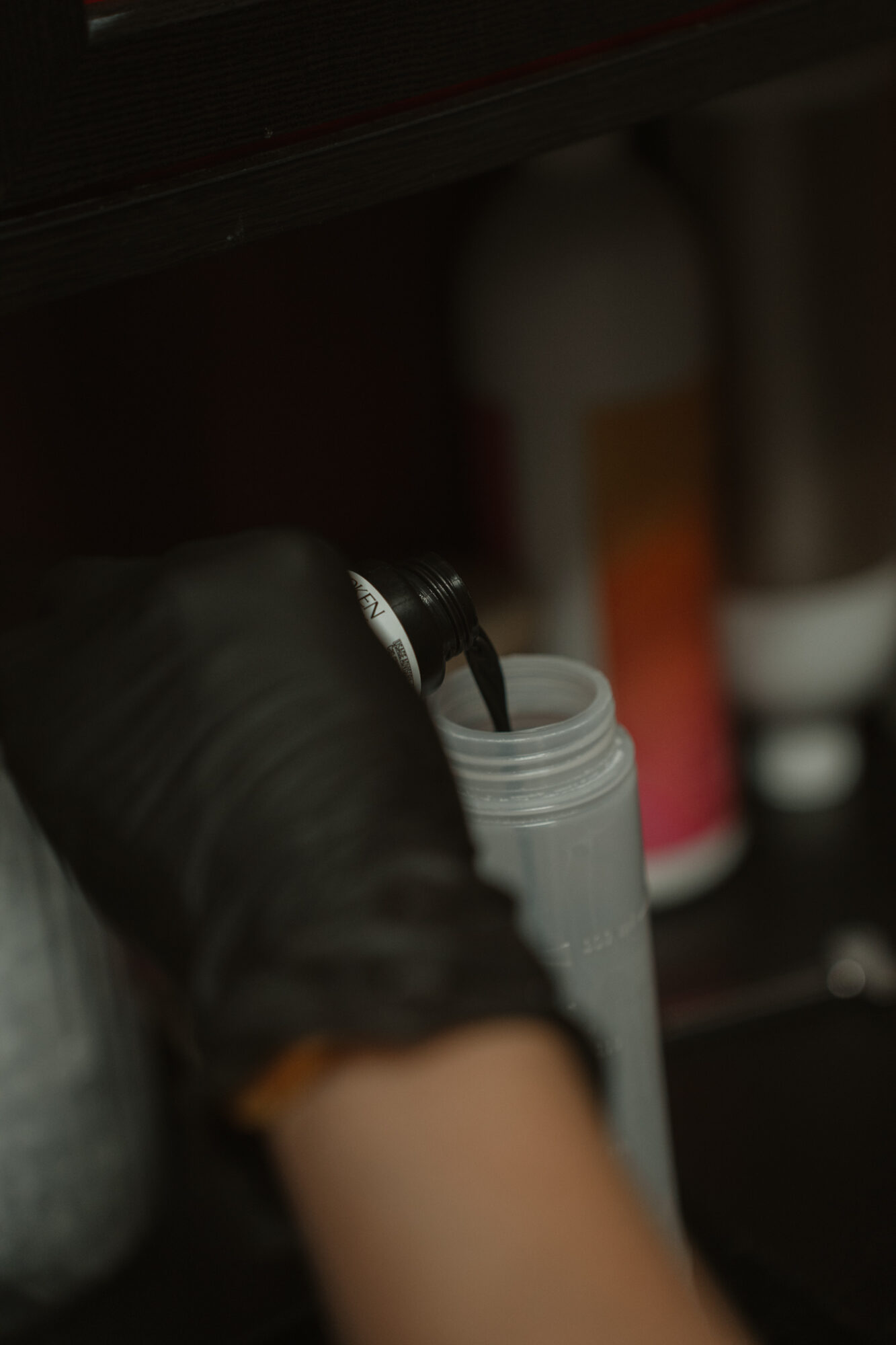 Person's arm and hand pouring liquid from a black bottle into a gray container, with blurred background objects.
