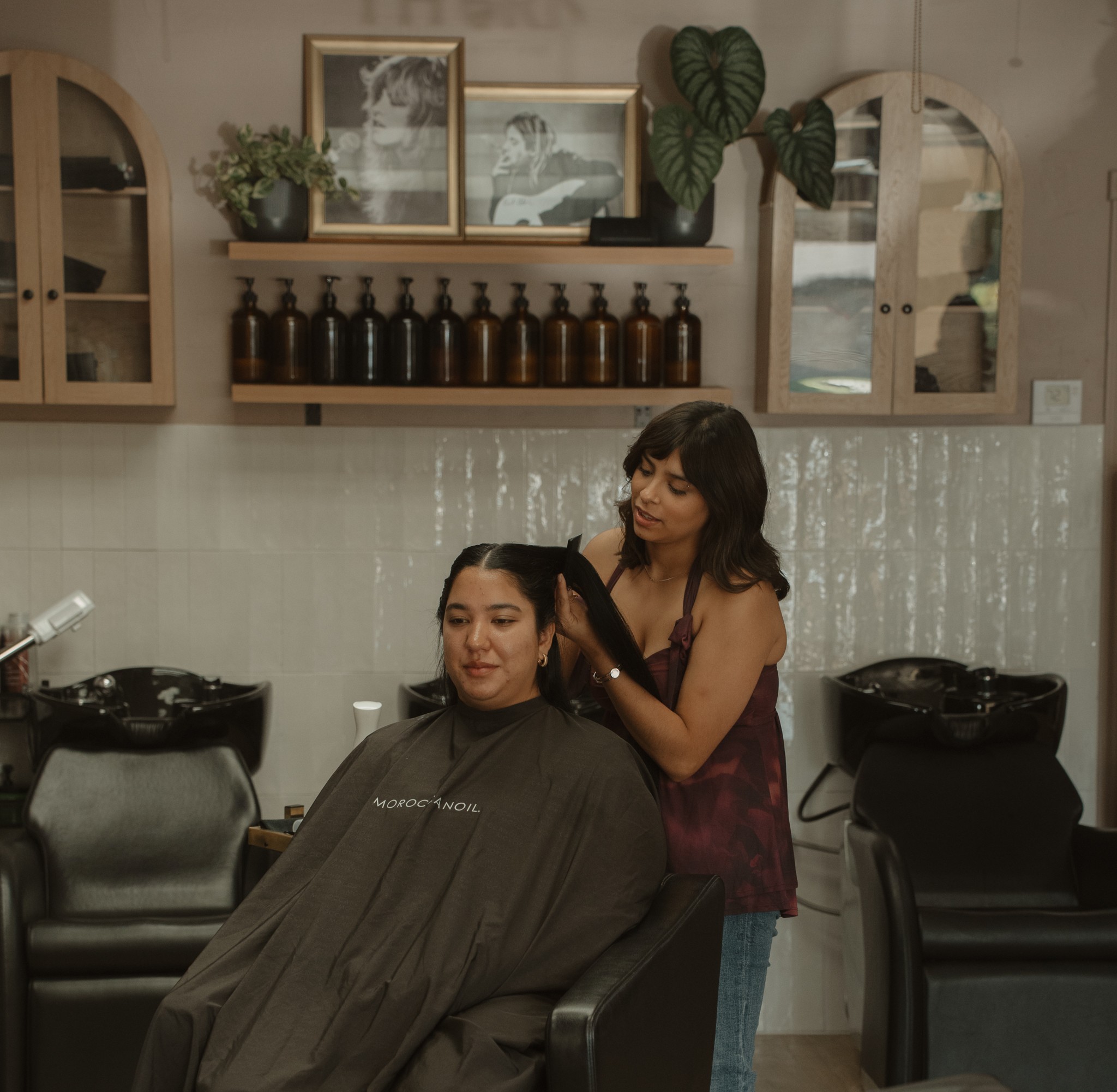 A woman gets her hair styled by a hairstylist in a salon with framed pictures and plants on the wall.