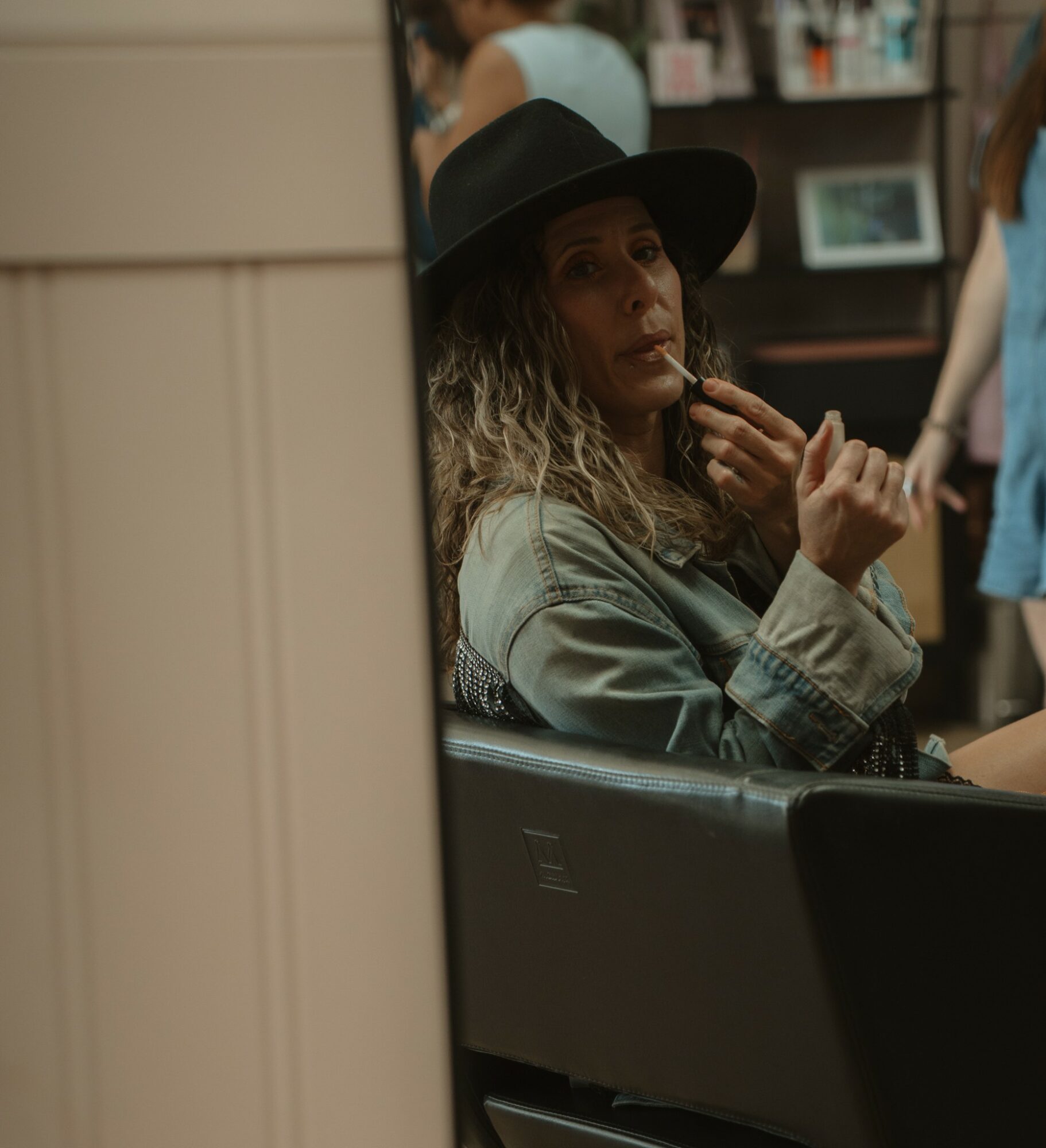 Woman with curly hair and hat sitting in a room, holding a cigarette, with books and a picture frame in the background.