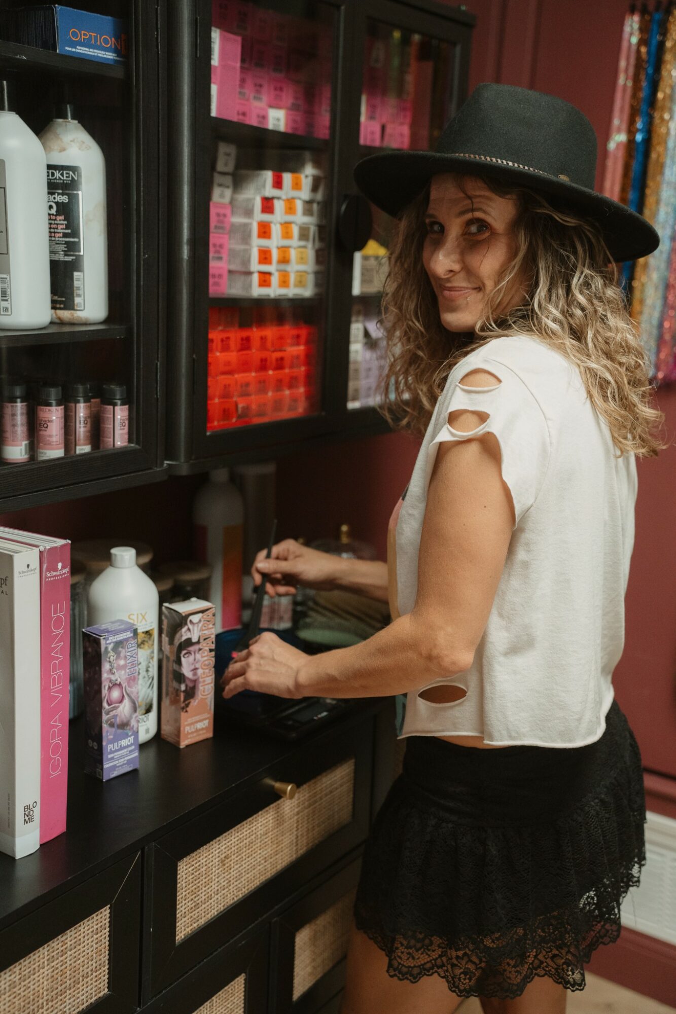 Woman with curly hair wearing a black hat and white top shopping for nail polish in store.