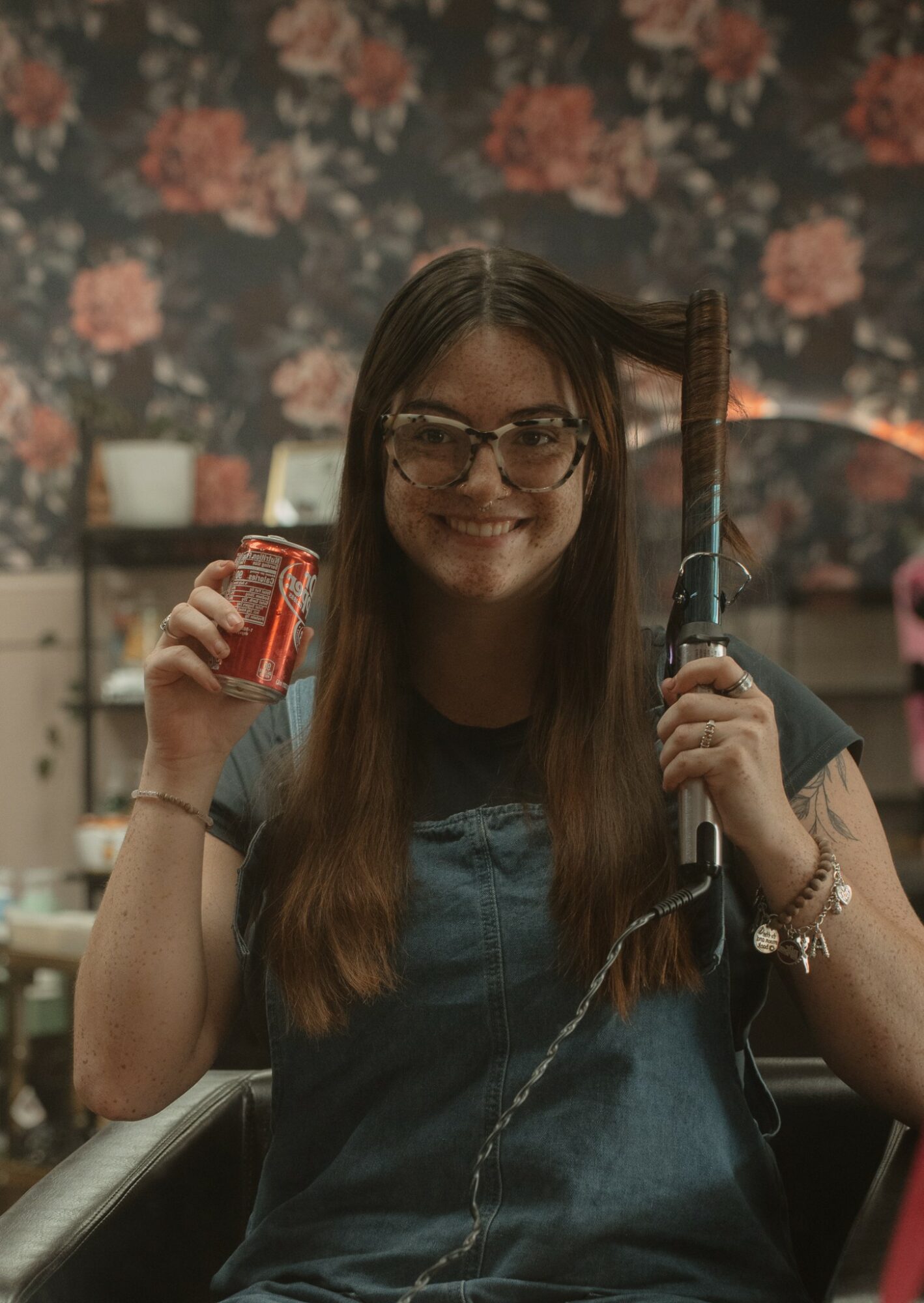 Young woman with glasses smiling, holding a soda can in one hand and a curling iron in the other, sitting indoors.