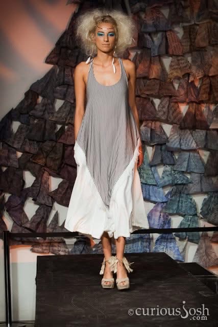 Model with curly hair in a layered dress on runway against a textured background.