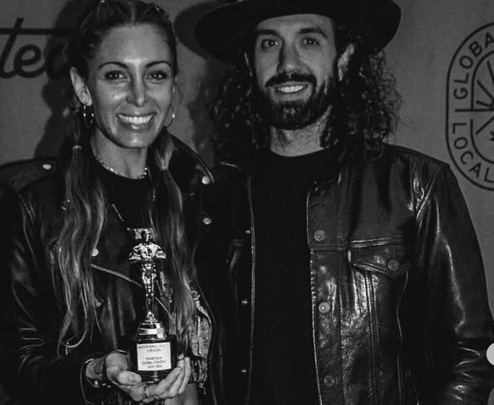 Two people smiling, one holding an award, both wearing leather jackets, in front of a backdrop with text and logos.