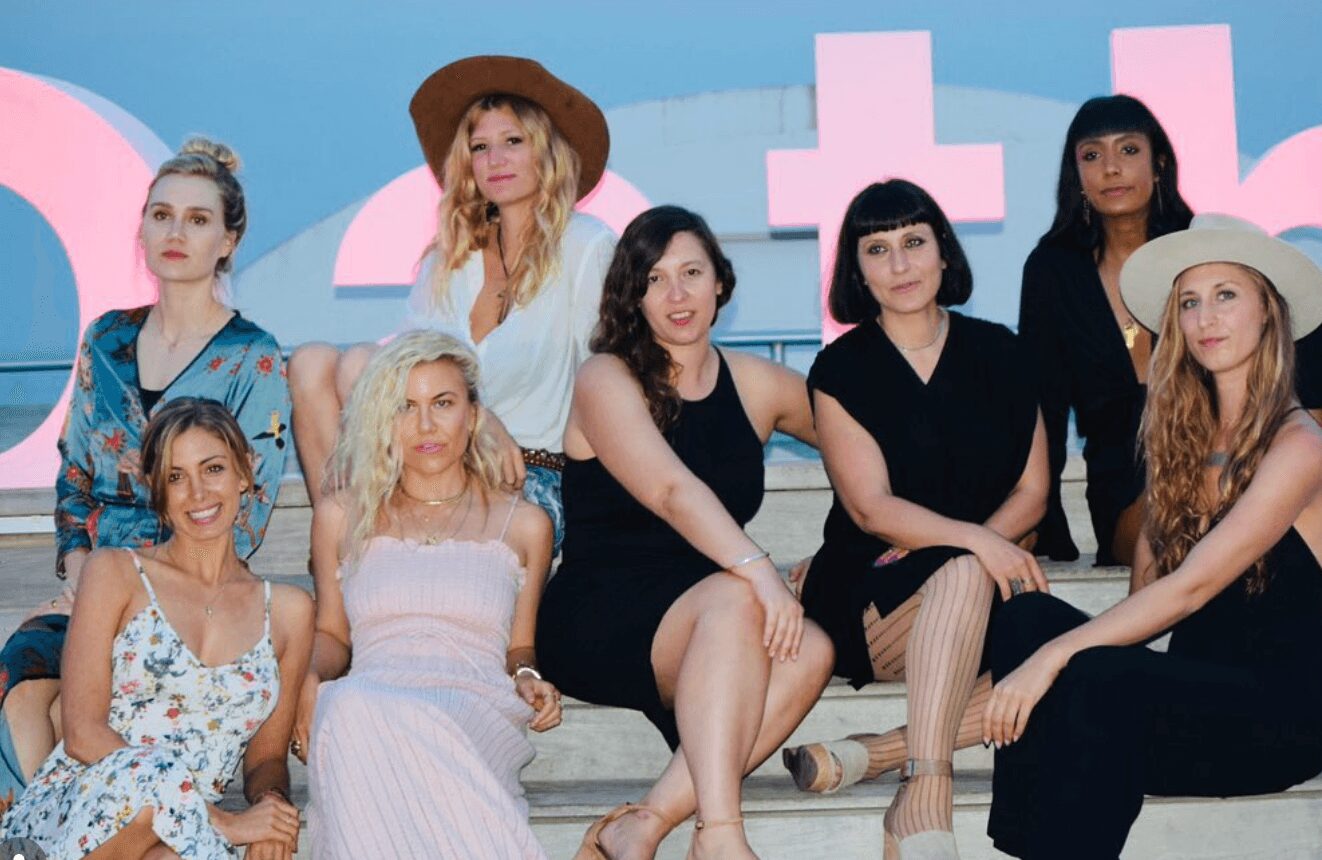Group of nine women sitting and standing on a stage with pink and blue background, some wearing hats and dresses.