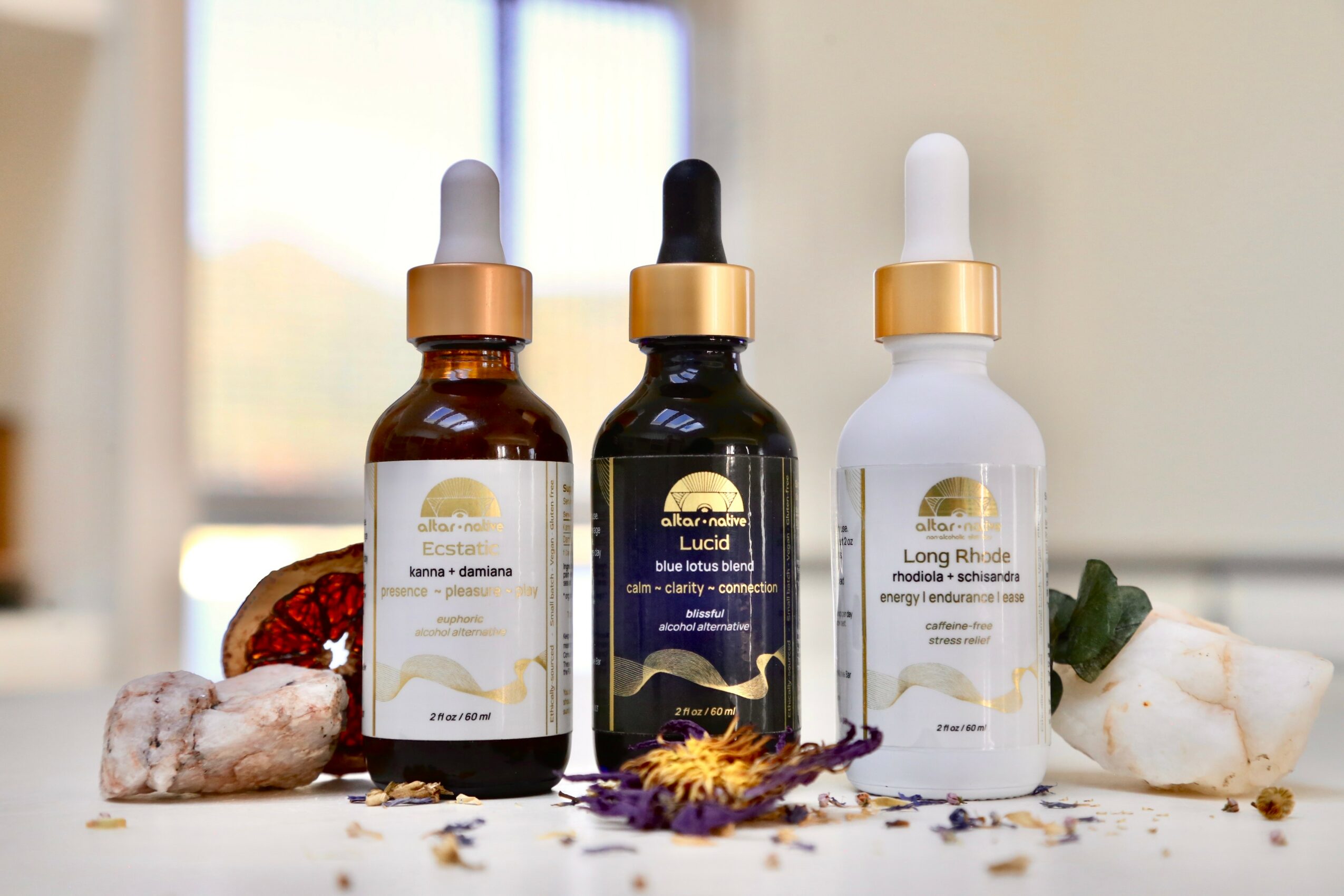 Three skincare serum bottles with dropper caps on a surface, surrounded by rocks and dried flowers, in a bright room.
