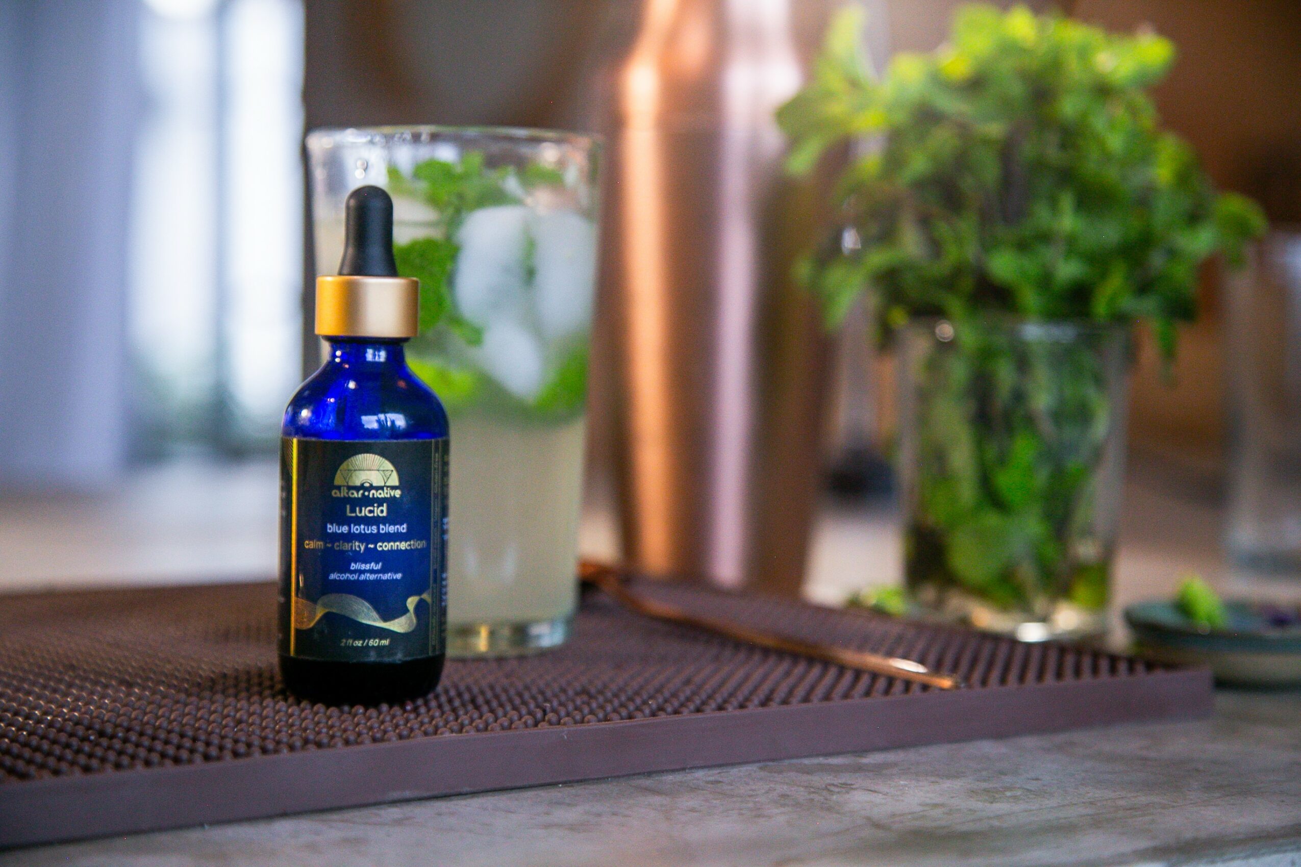 Blue glass dropper bottle with a gold cap on a table, with a glass of water and a potted plant in the background.