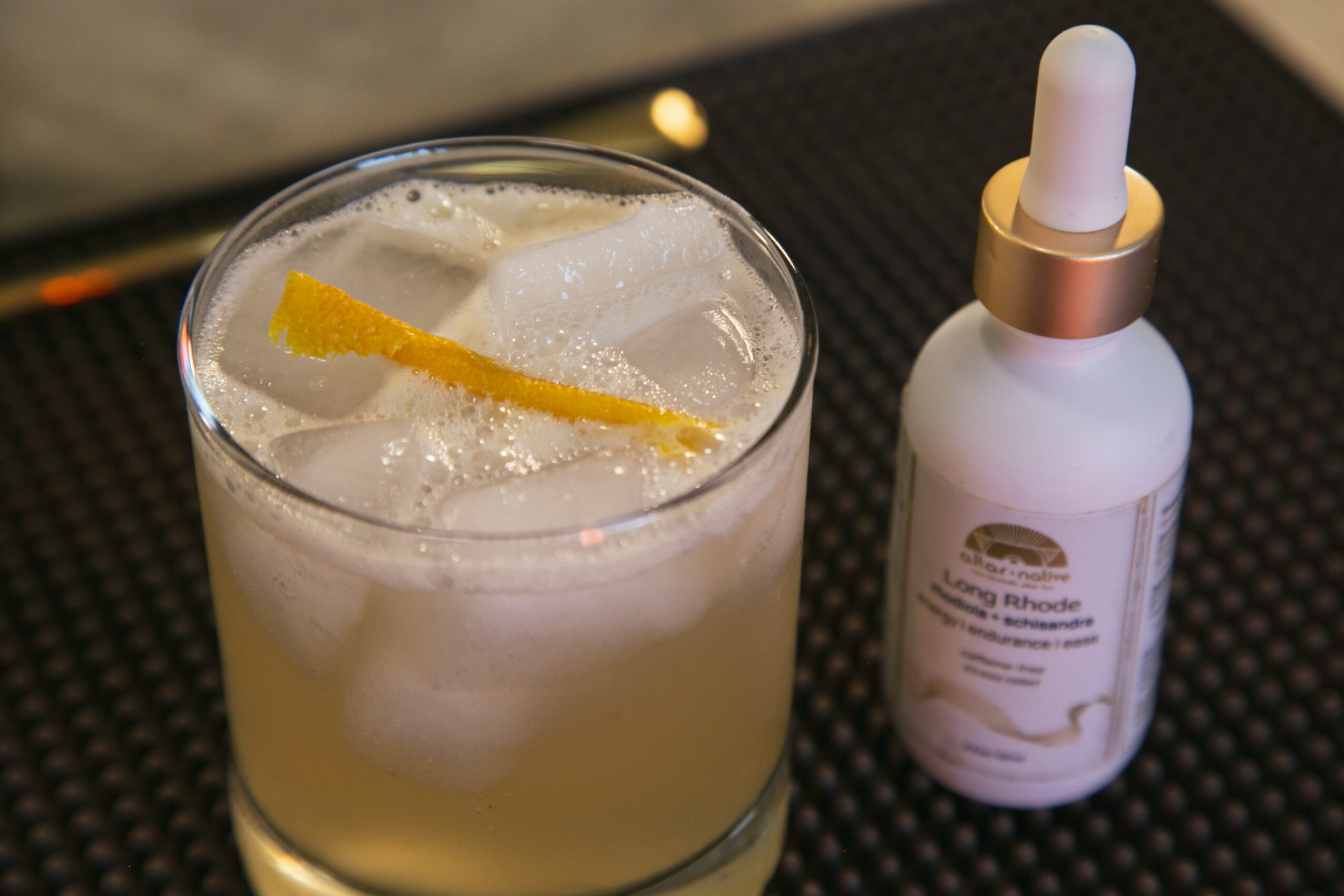 A glass with a light-colored beverage and ice, next to a white dropper bottle with a gold band, on a black surface.