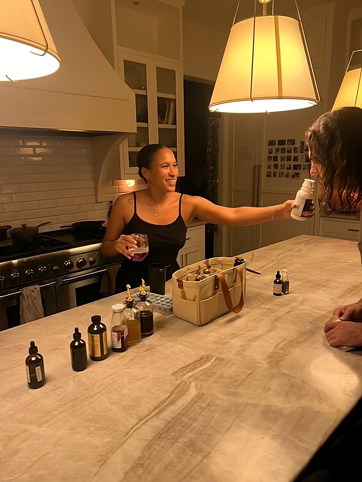 Woman serving drinks at a kitchen counter with bottles and a bag, illuminated by hanging lamps.