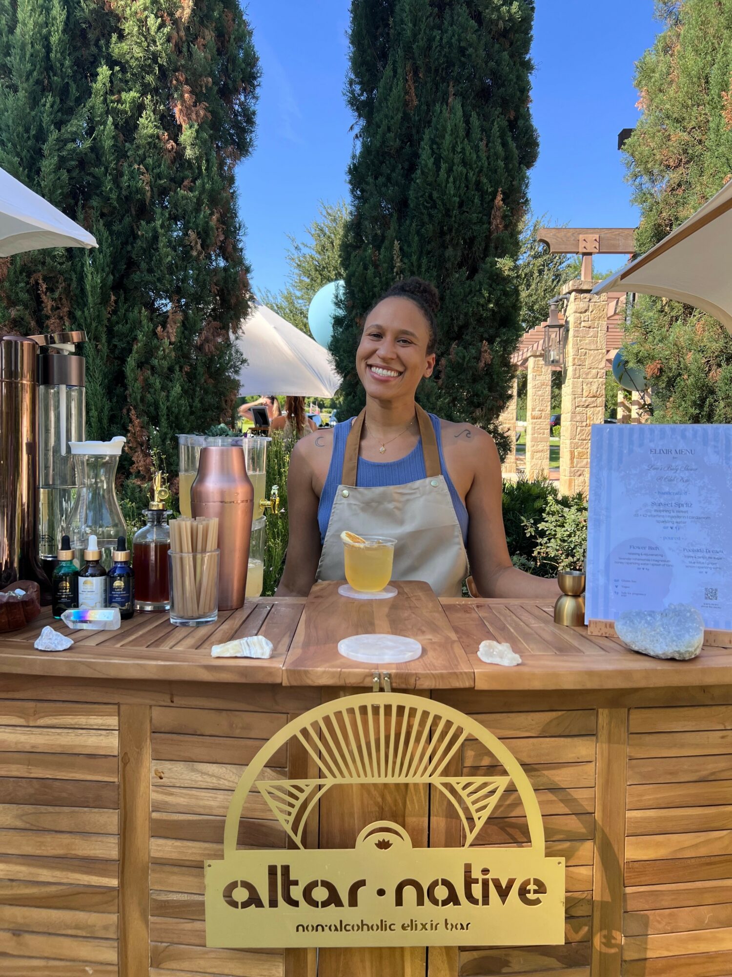 Smiling woman standing behind a wooden outdoor bar with trees and umbrellas in background.