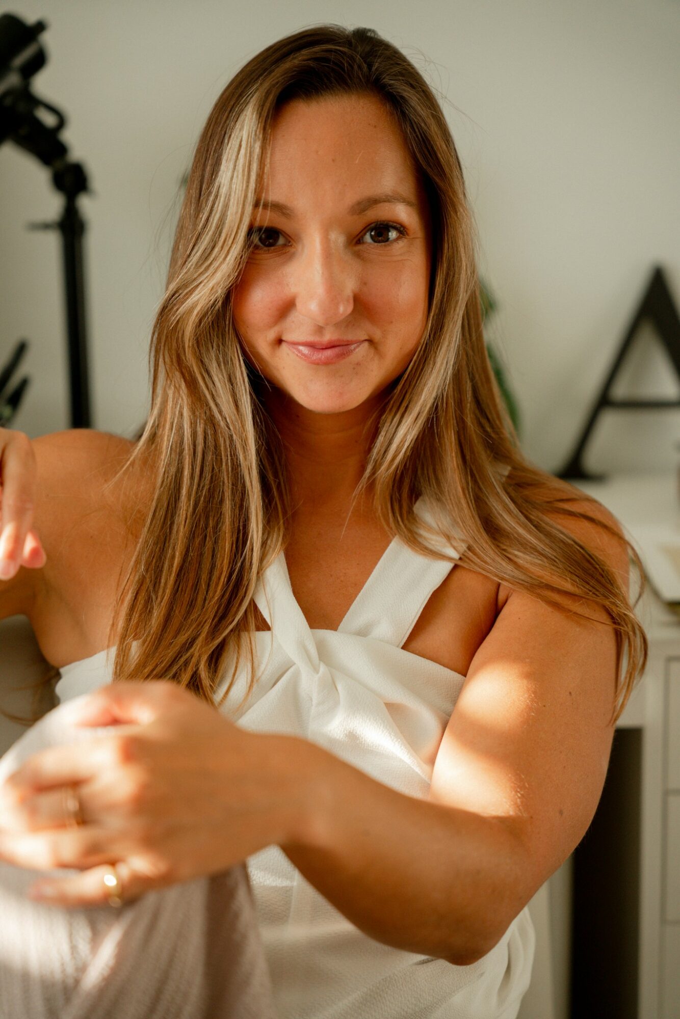 Young woman with long hair smiling, wearing a white dress, in a well-lit room with decorative elements.