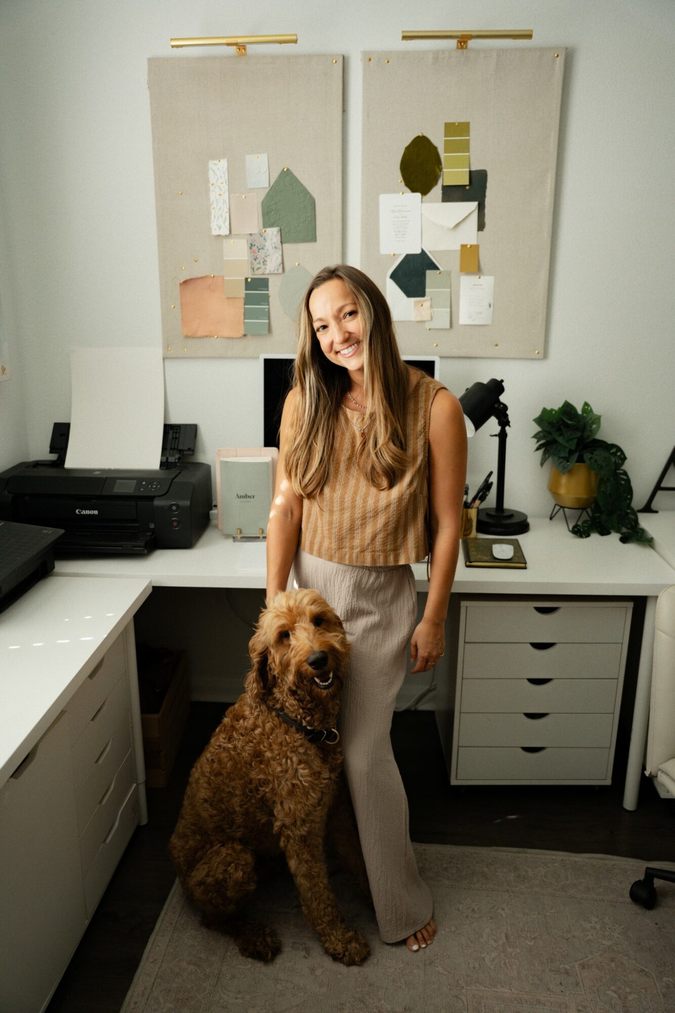 Woman with long hair standing in an office with a dog, two abstract wall hangings, and office equipment.