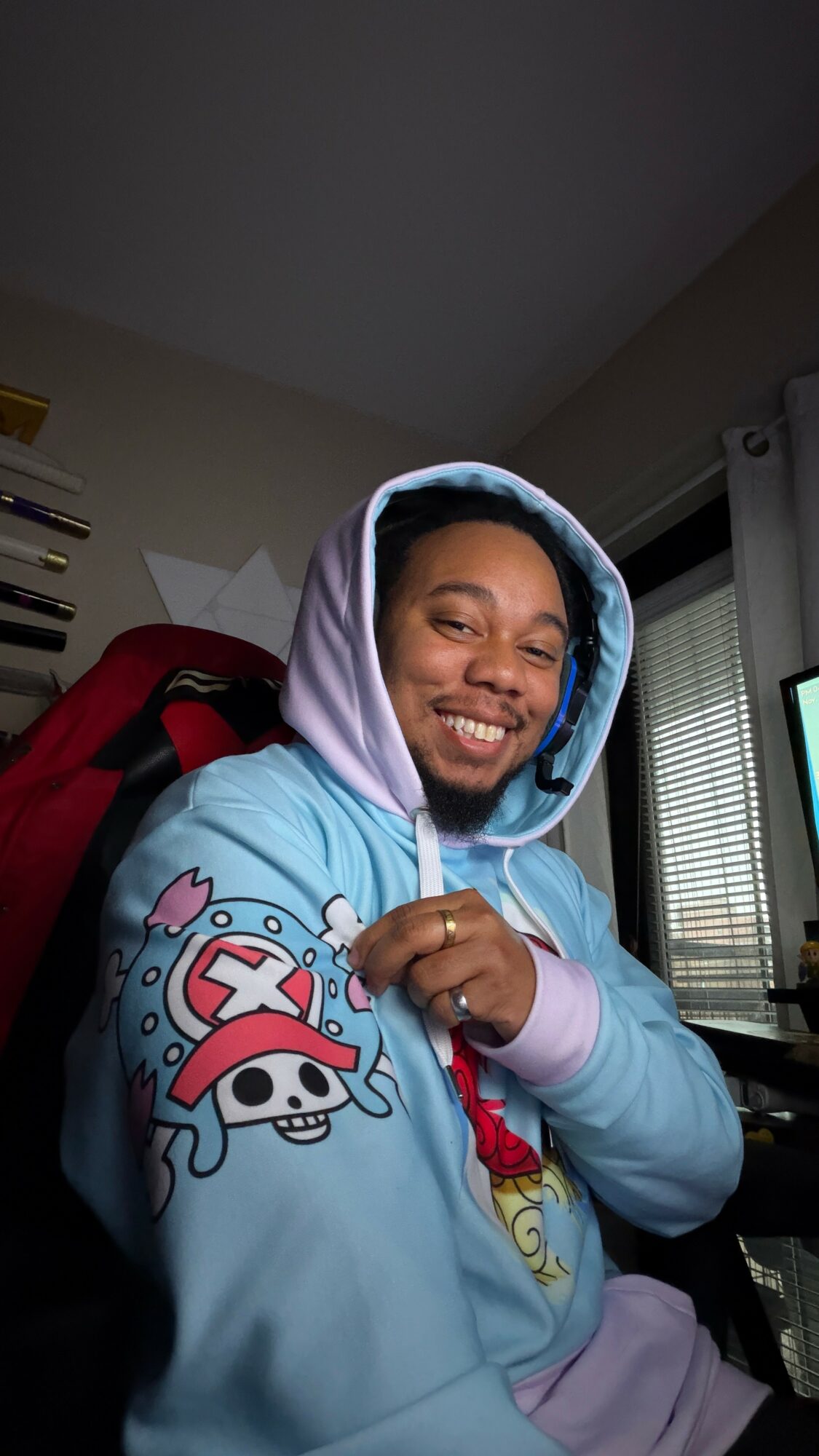 Person smiling, wearing a hoodie with cartoon characters, sitting in a room with a computer and window blinds