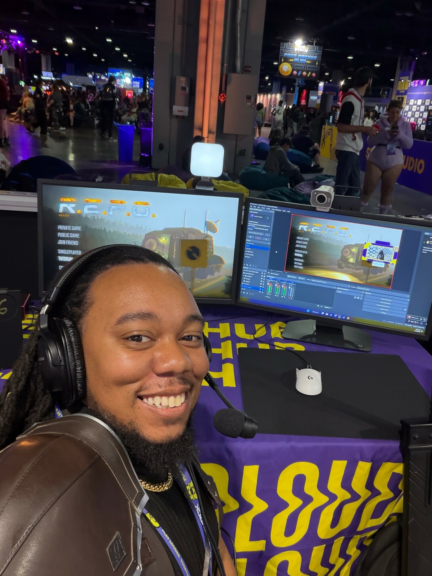 Person with headphones smiling in front of two computer monitors at an indoor event, with people and lights in the background.