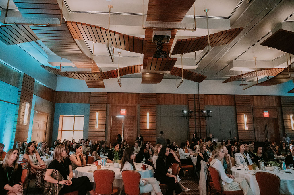 Large banquet hall filled with people seated at round tables, decorated with elegant lighting and ceiling design.