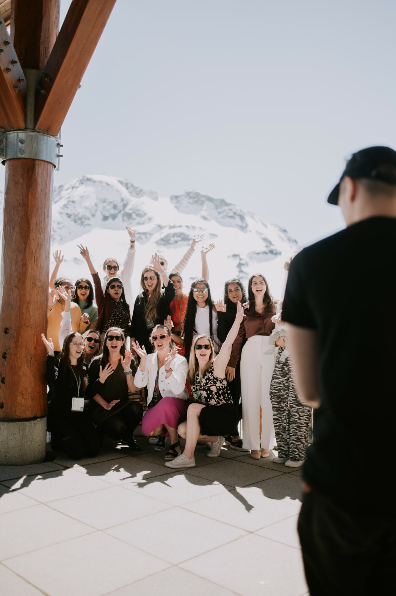 Group of people posing outdoors with snowy mountains in background, some raising hands, some taking photos, person in foreground facing away.