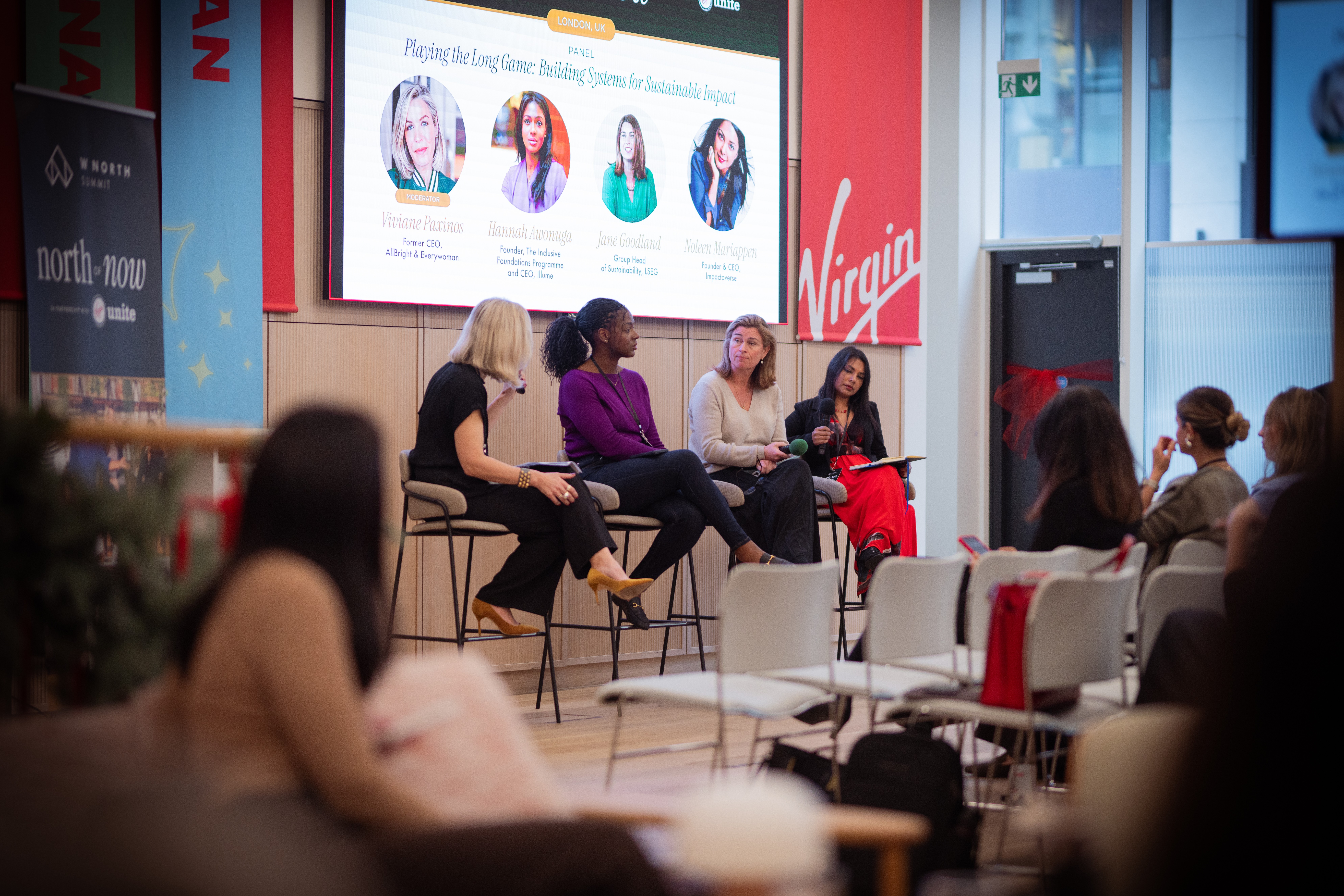 Panel of four women speaking on stage in front of an audience, with a large screen behind them displaying images and text.
