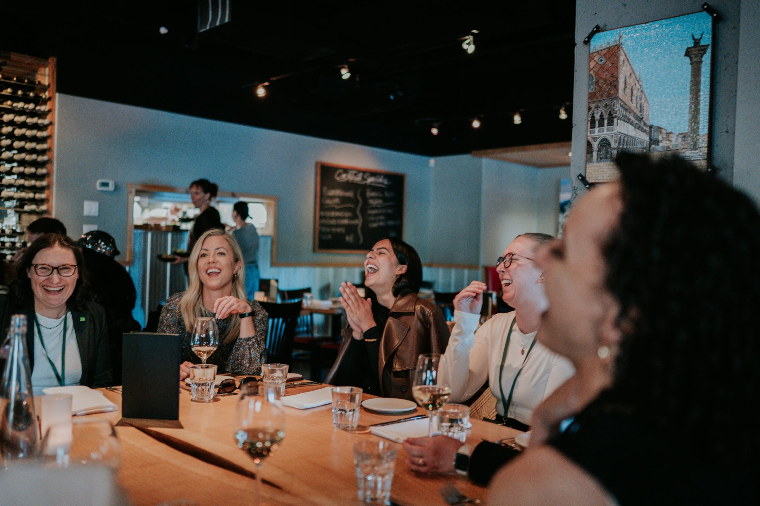 Group of people laughing and enjoying drinks at a bar or restaurant, with a chalkboard menu in the background.