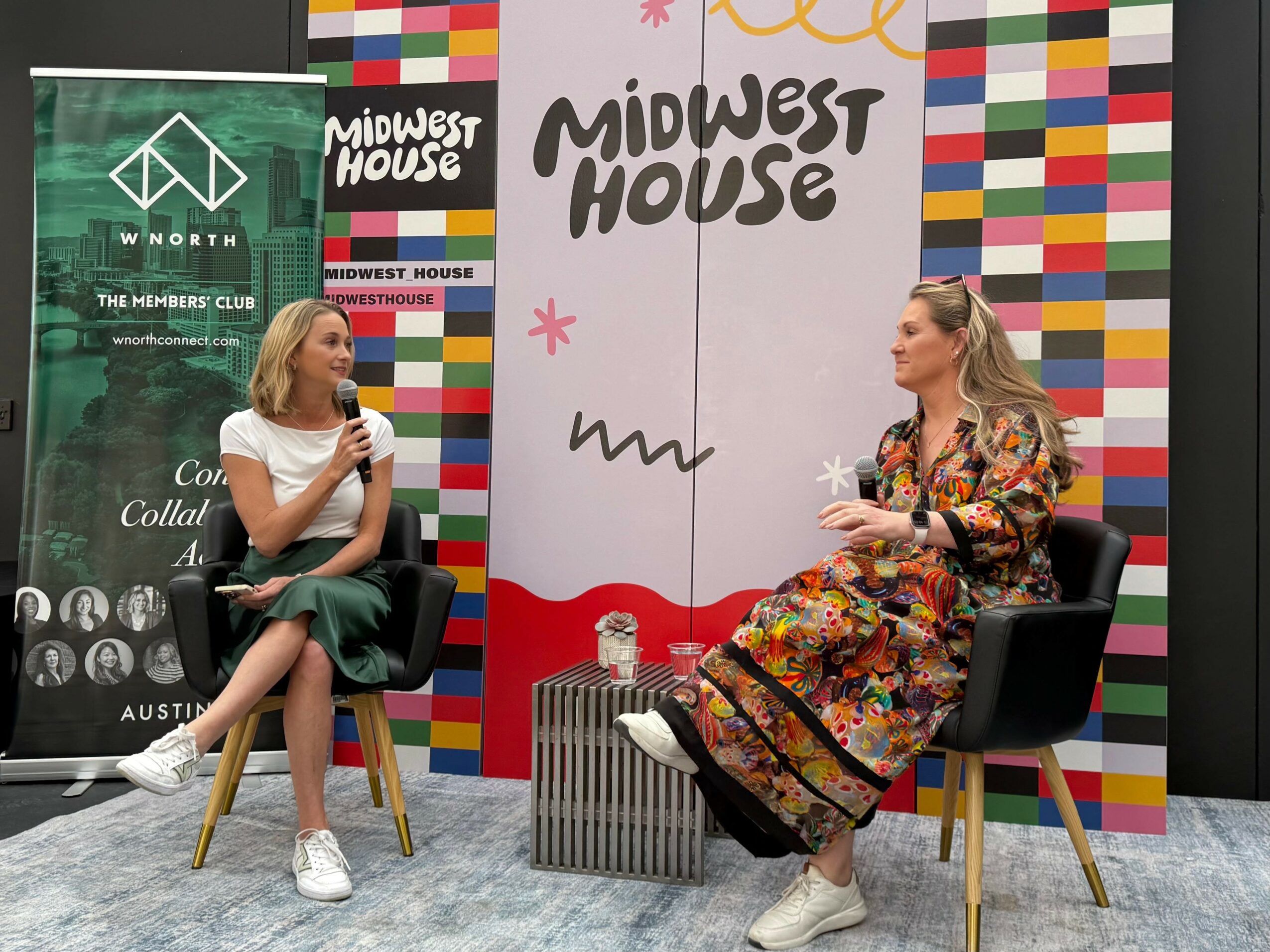 Two women seated on stage, engaged in conversation, with colorful backdrop and banners behind them.