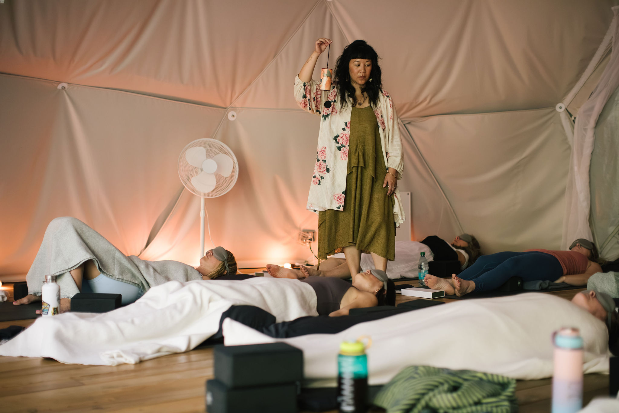 Person standing on stage with people lying down, in a tent-like setting with soft lighting, fan, and blankets.