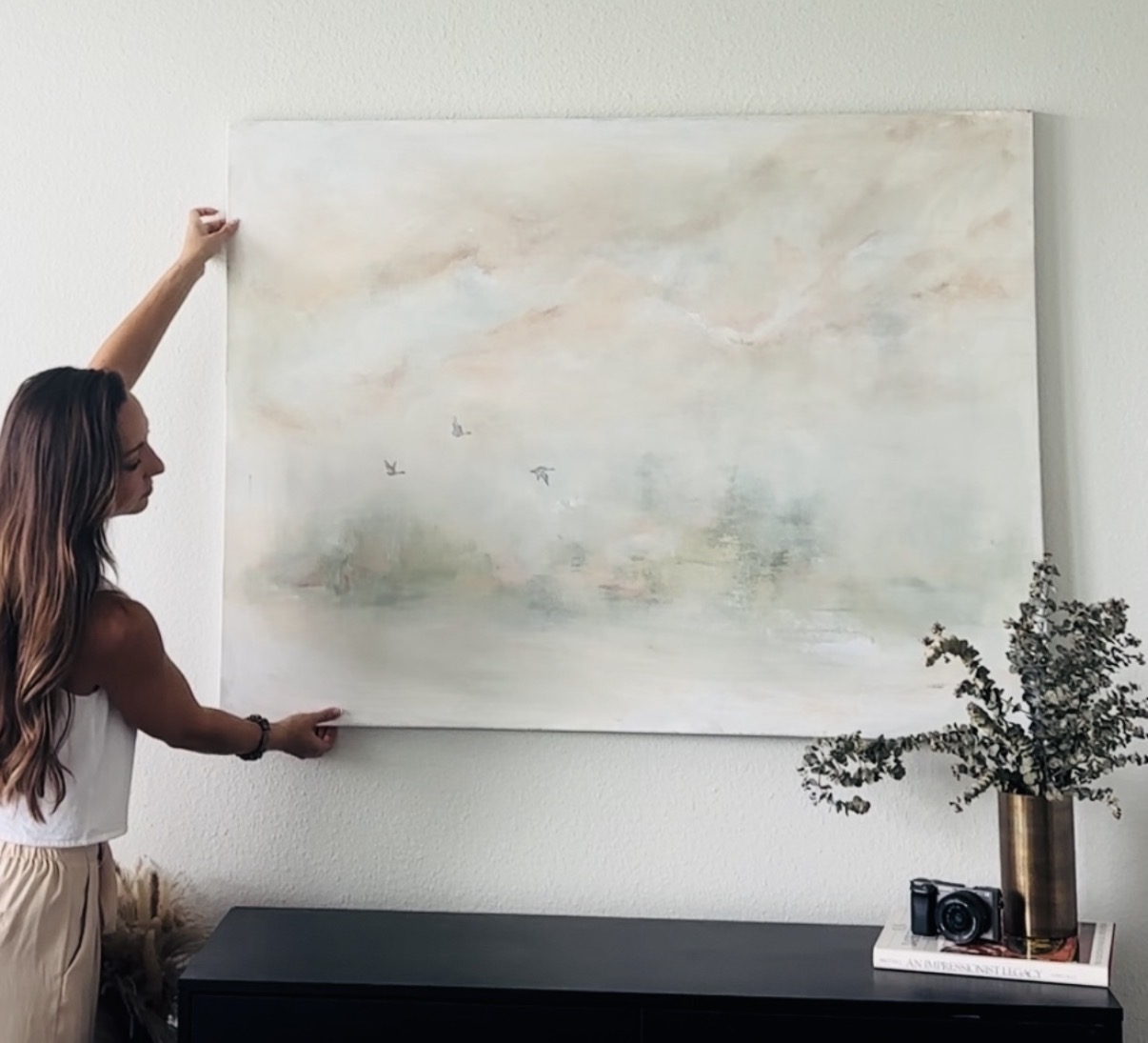 Woman hanging a large abstract painting on a white wall, with a black console table and a potted plant nearby.