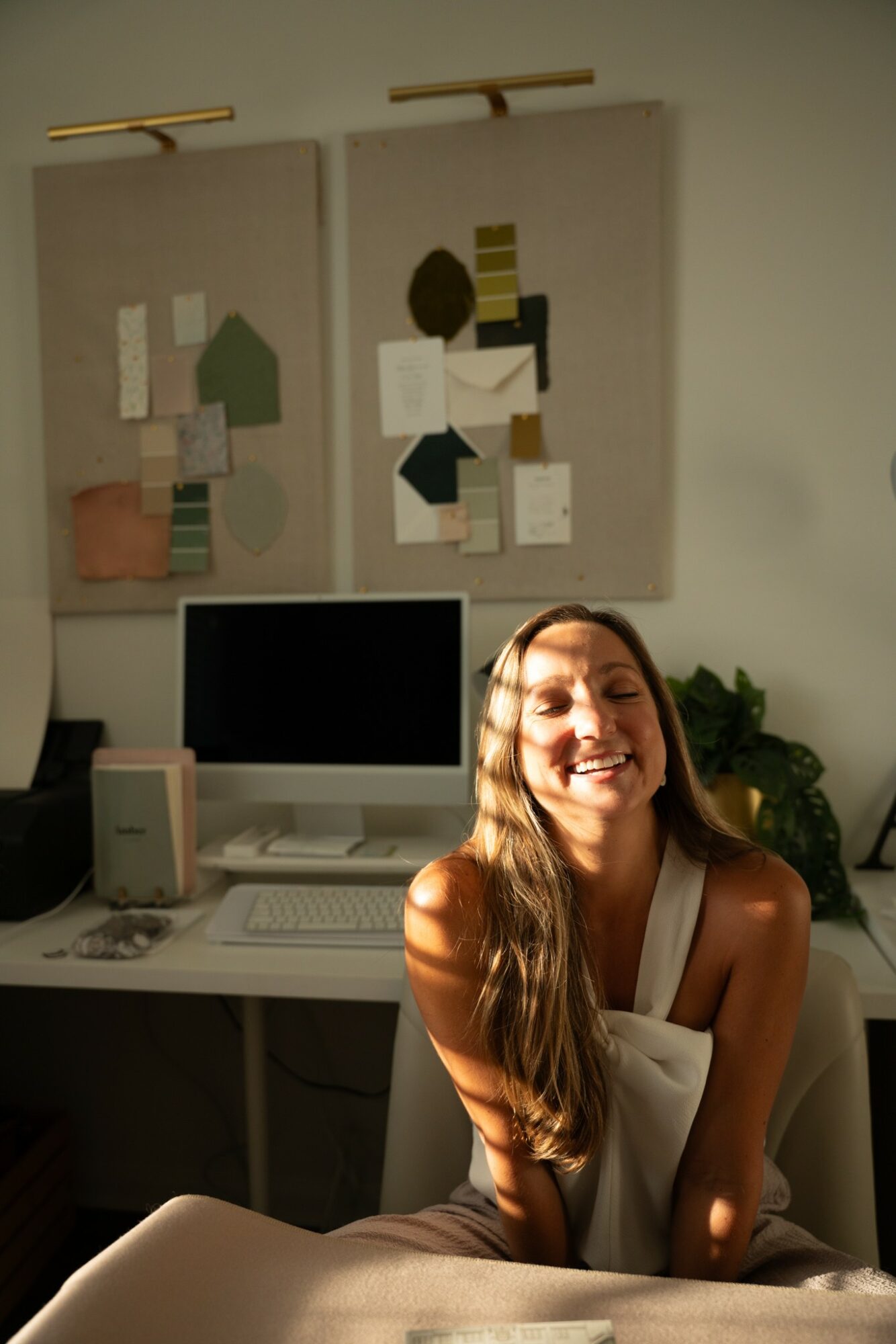 Smiling woman with long hair sitting in front of a desk with a computer, two abstract art pieces on the wall behind her.