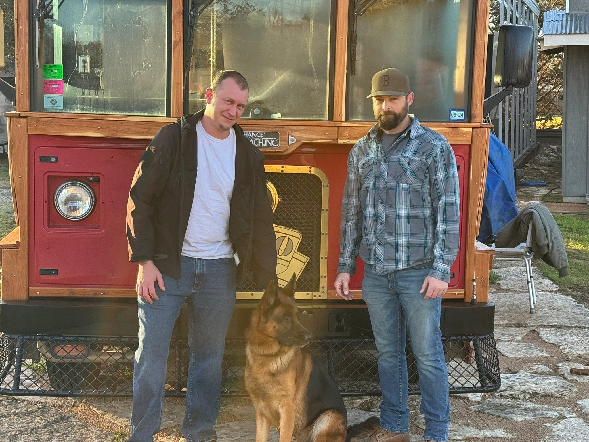 Two men stand outdoors with a German Shepherd dog in front of a food truck, one wearing a black jacket and the other a plaid shirt.