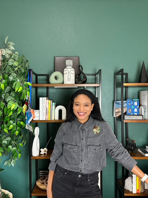 Smiling woman with long dark hair in gray jacket standing in front of green wall and bookshelves.