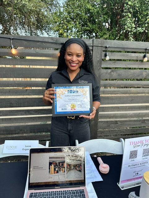 Woman holding a framed certificate, standing outdoors near a wooden fence, with a table displaying a laptop and papers nearby.