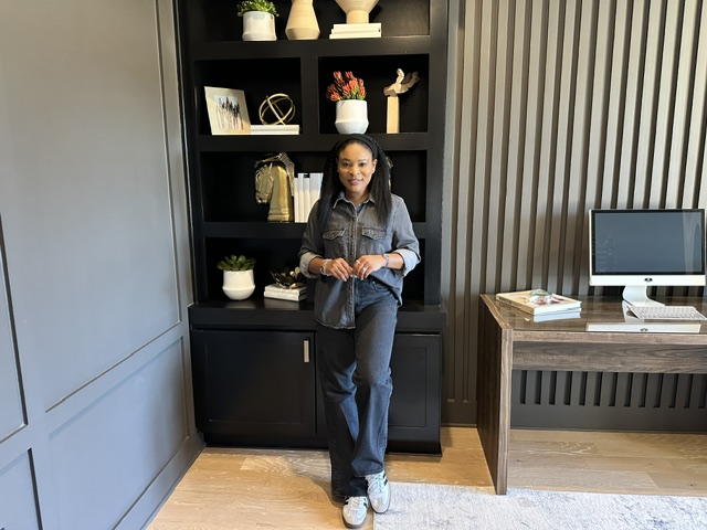 Girl standing in front of a black bookshelf, holding a bag, with a desk and computer to the right, in a modern room.