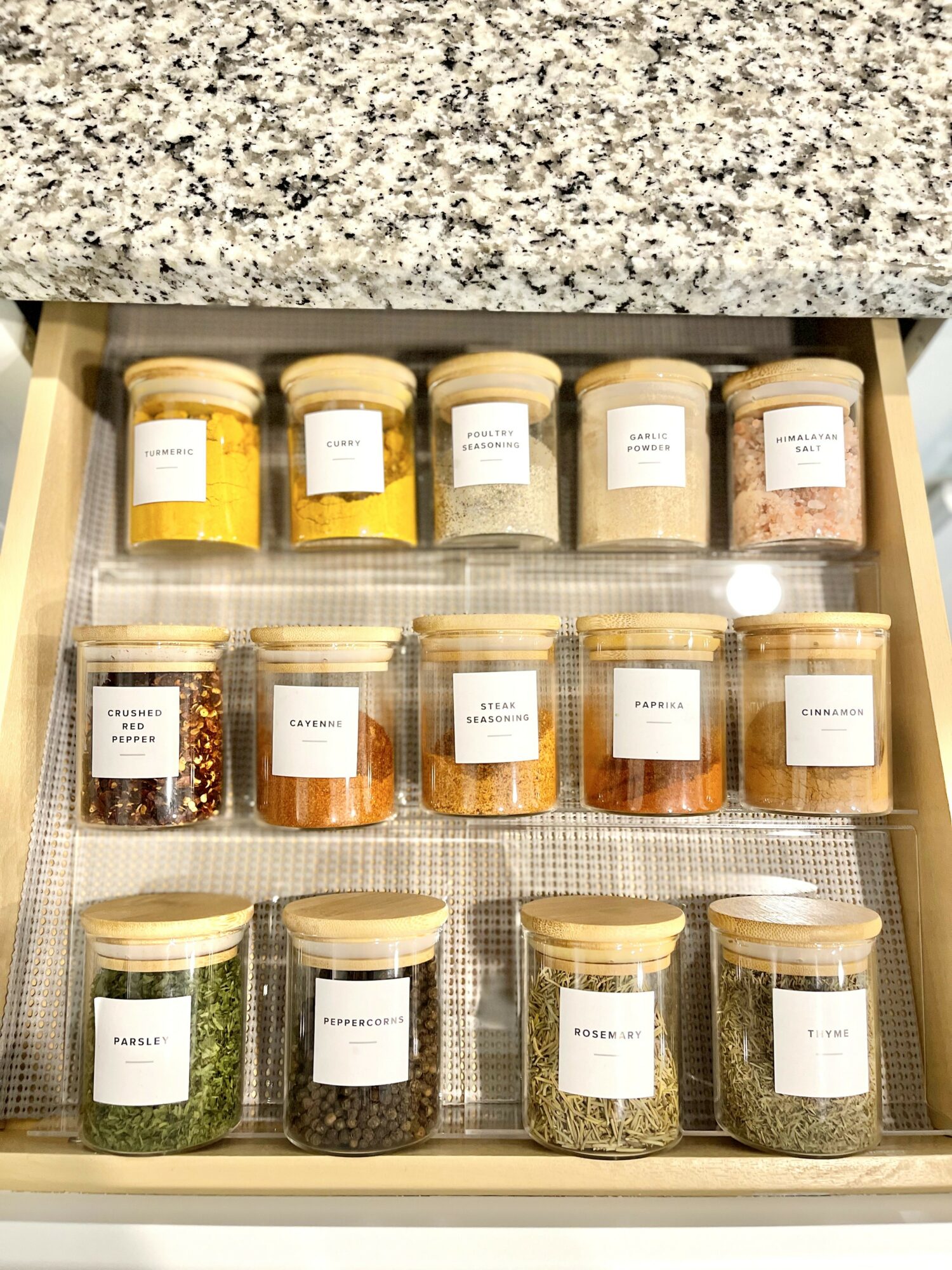 Open drawer with 12 jars of spices and herbs, organized in three rows on a textured surface.