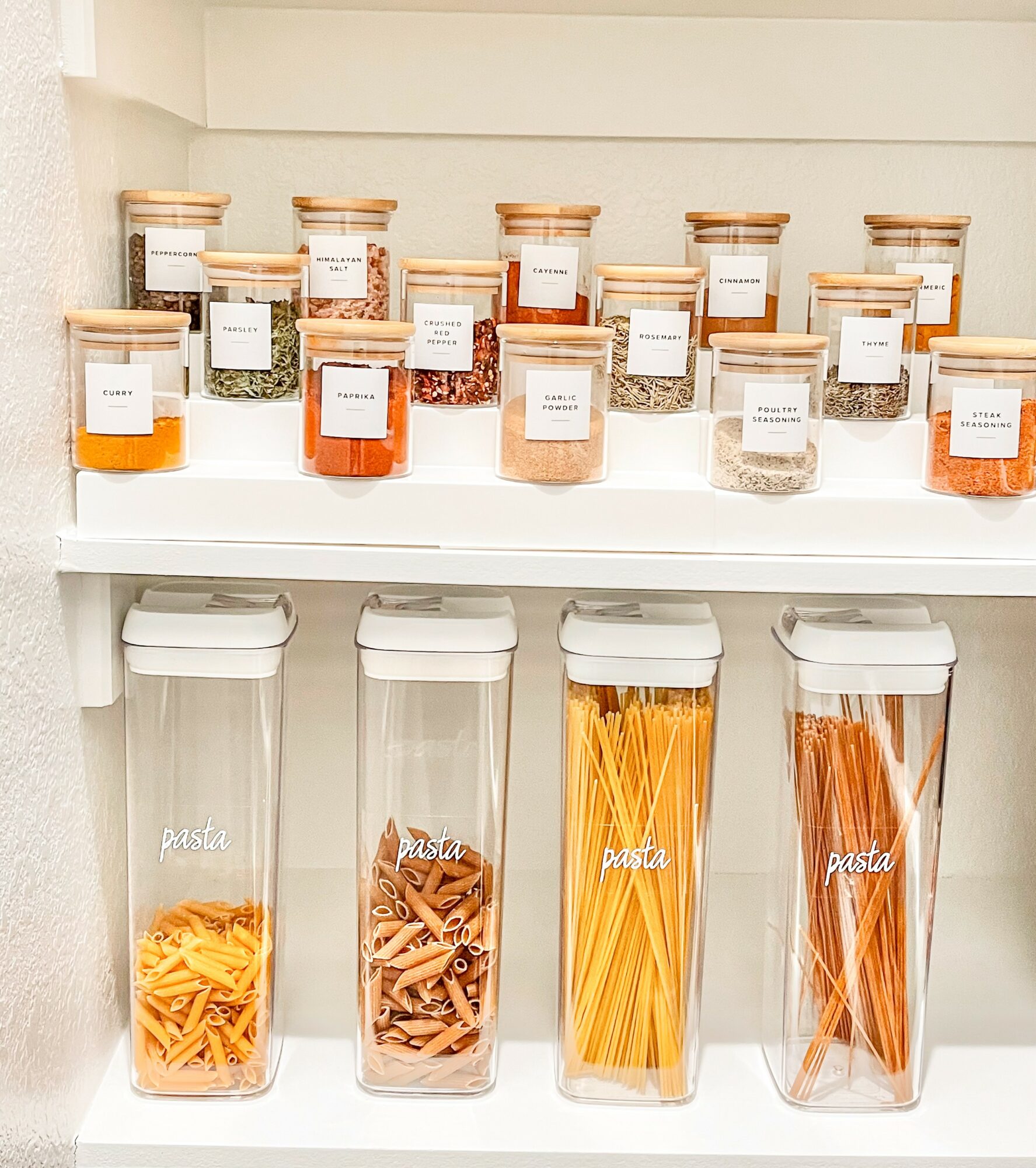 Shelf with glass jars of spices on top and pasta containers below, in a kitchen setting.