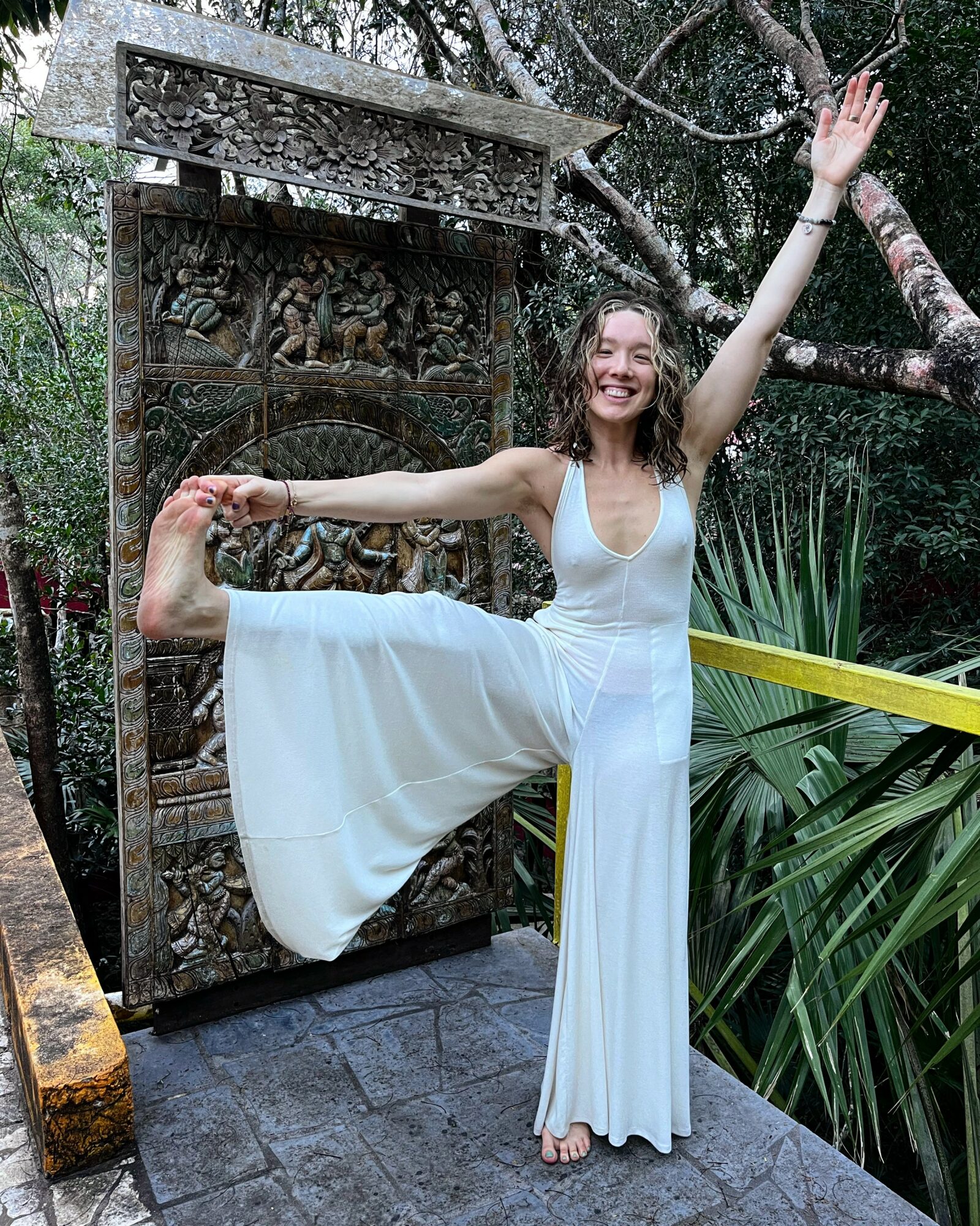 Woman in white dress balancing on one leg outdoors, smiling, with one arm raised and the other holding her foot, surrounded by greenery.