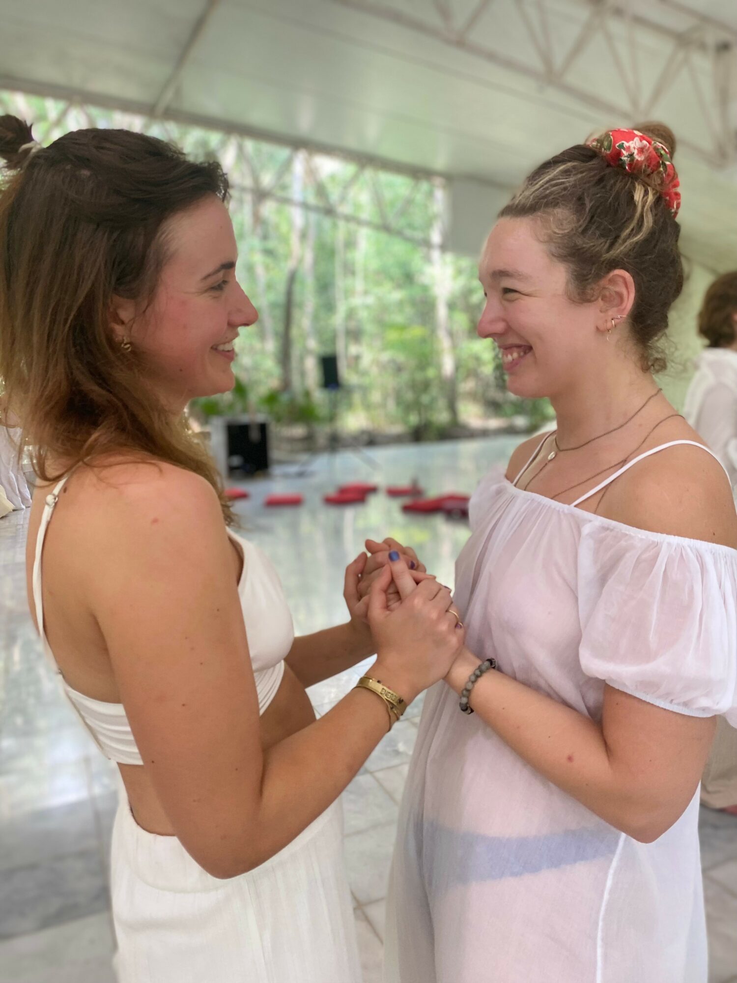 Two women smiling and holding hands indoors with a glass ceiling and greenery outside, wearing light-colored dresses.