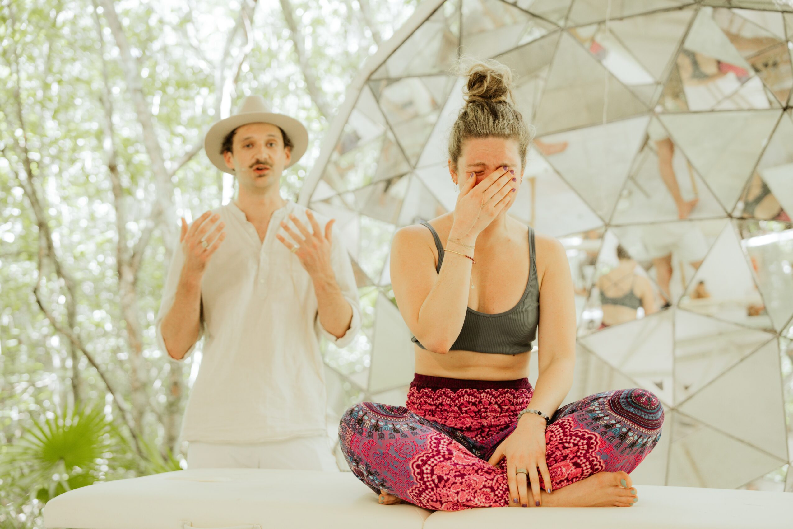 Two women outdoors, one covering her face, the other speaking, with a reflective geometric sculpture behind them.