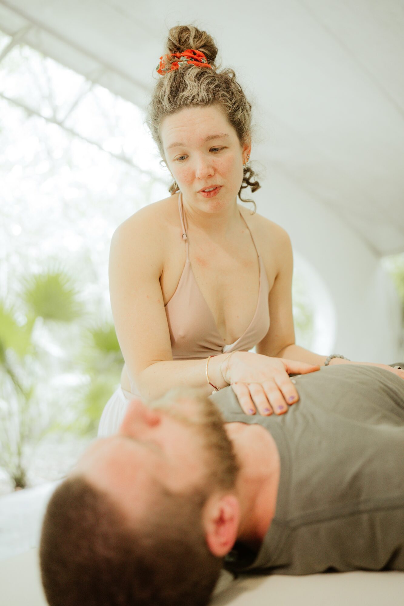 Young woman with curly hair and a red hair accessory leans over a man lying down, indoors with bright background.