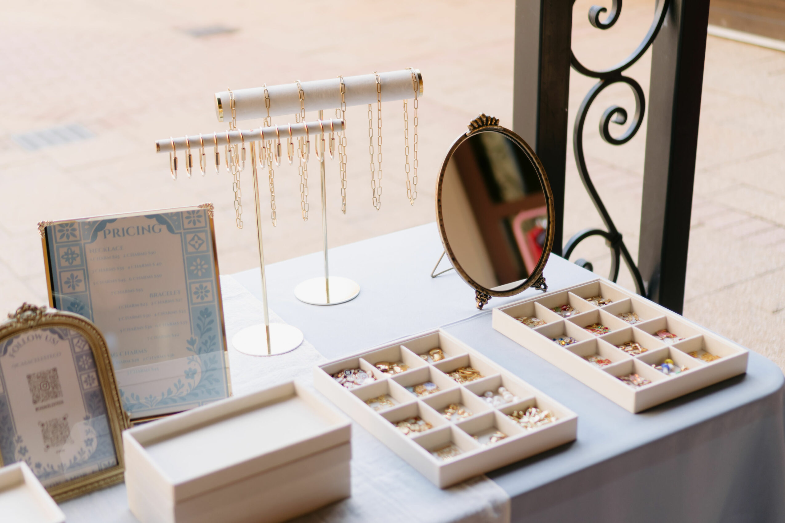 Jewelry display table with necklaces, rings, and a mirror, set outdoors on a sidewalk.
