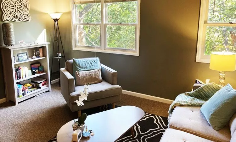 Living room with armchair, bookshelf, windows, and sofa with pillows, illuminated by lamps and natural light.