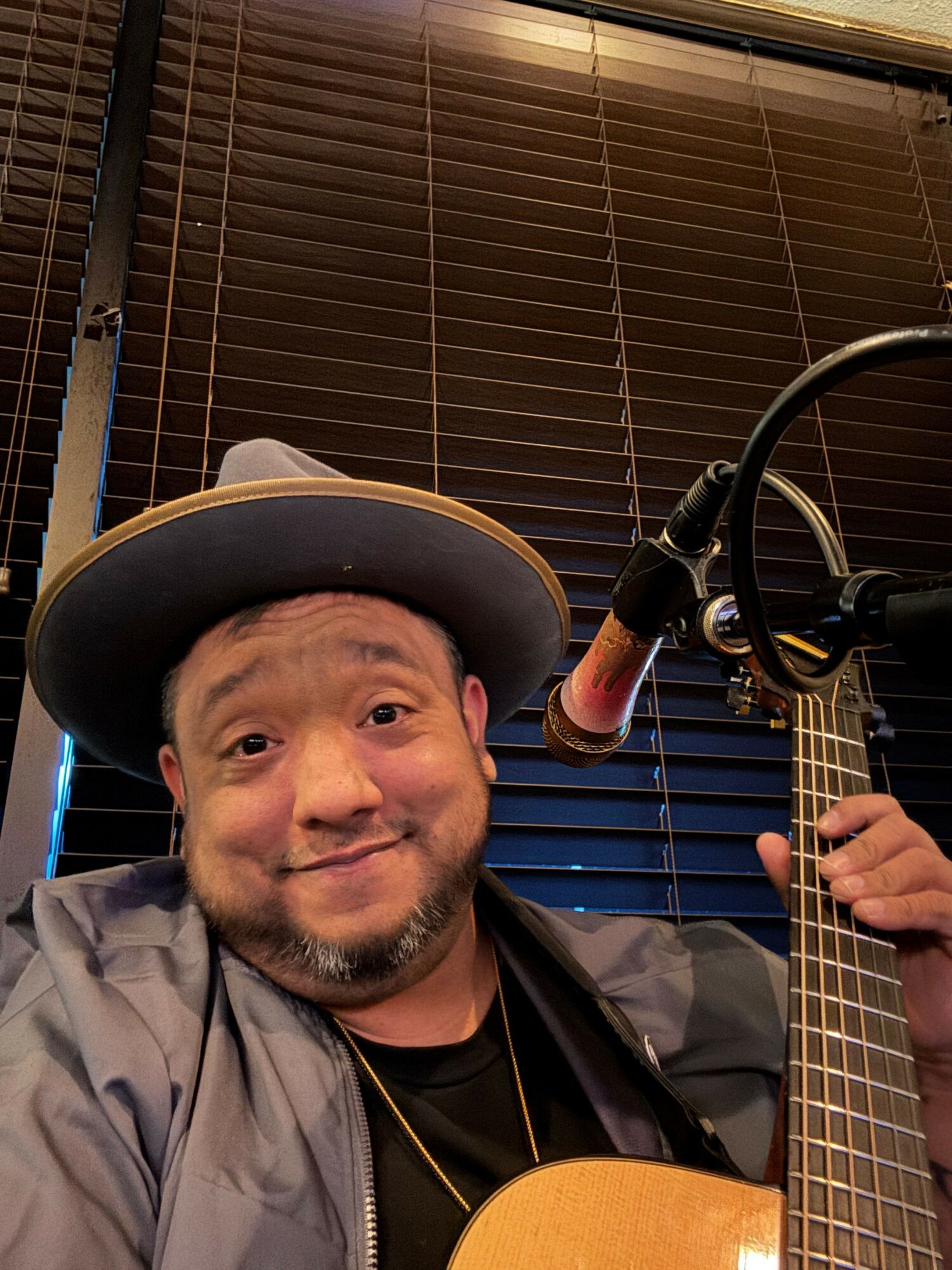 Person wearing a hat, playing guitar in a recording studio with a microphone, dark background, and wooden panels.