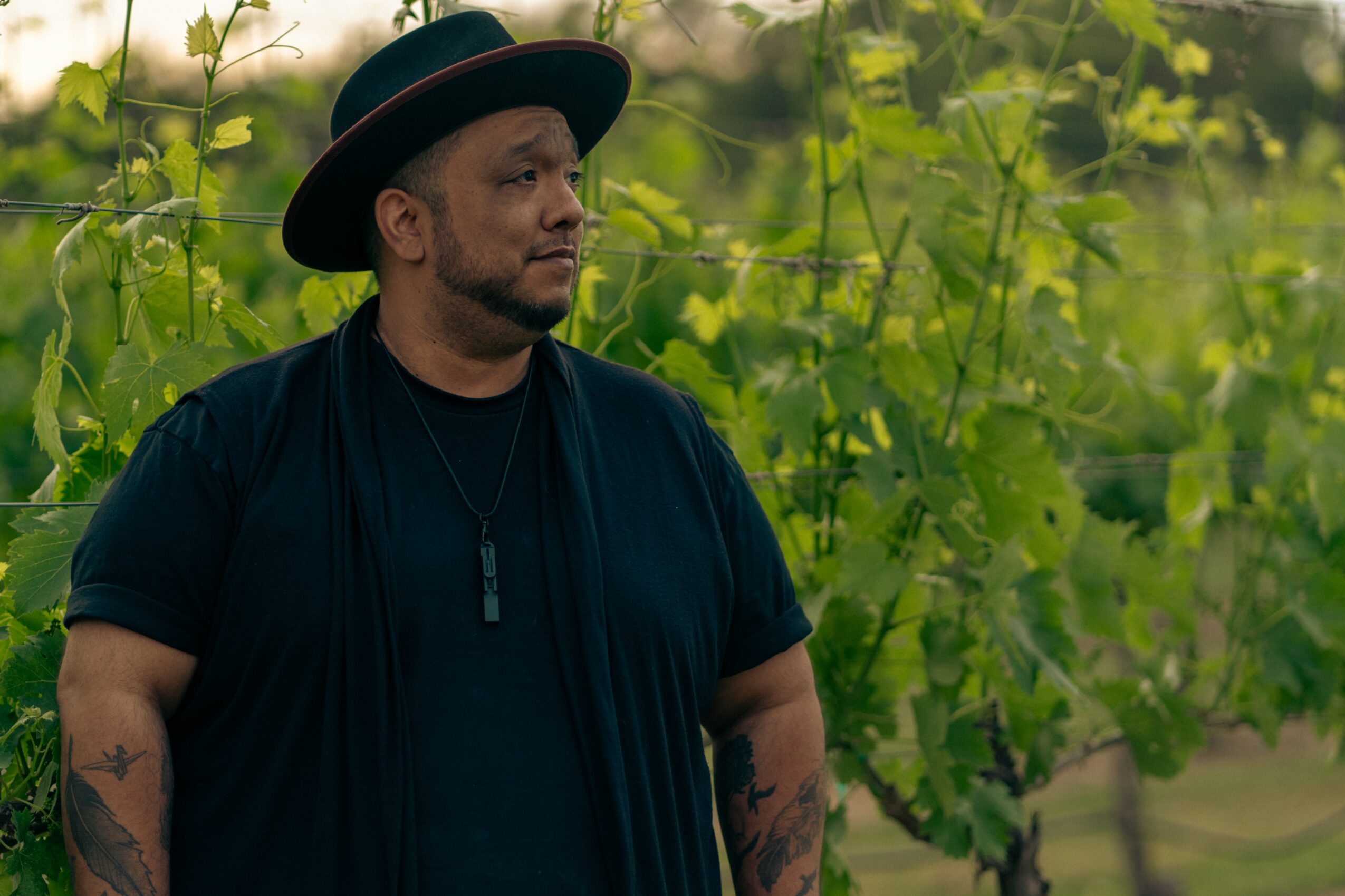 Man wearing a black hat and shirt stands outdoors among green foliage.