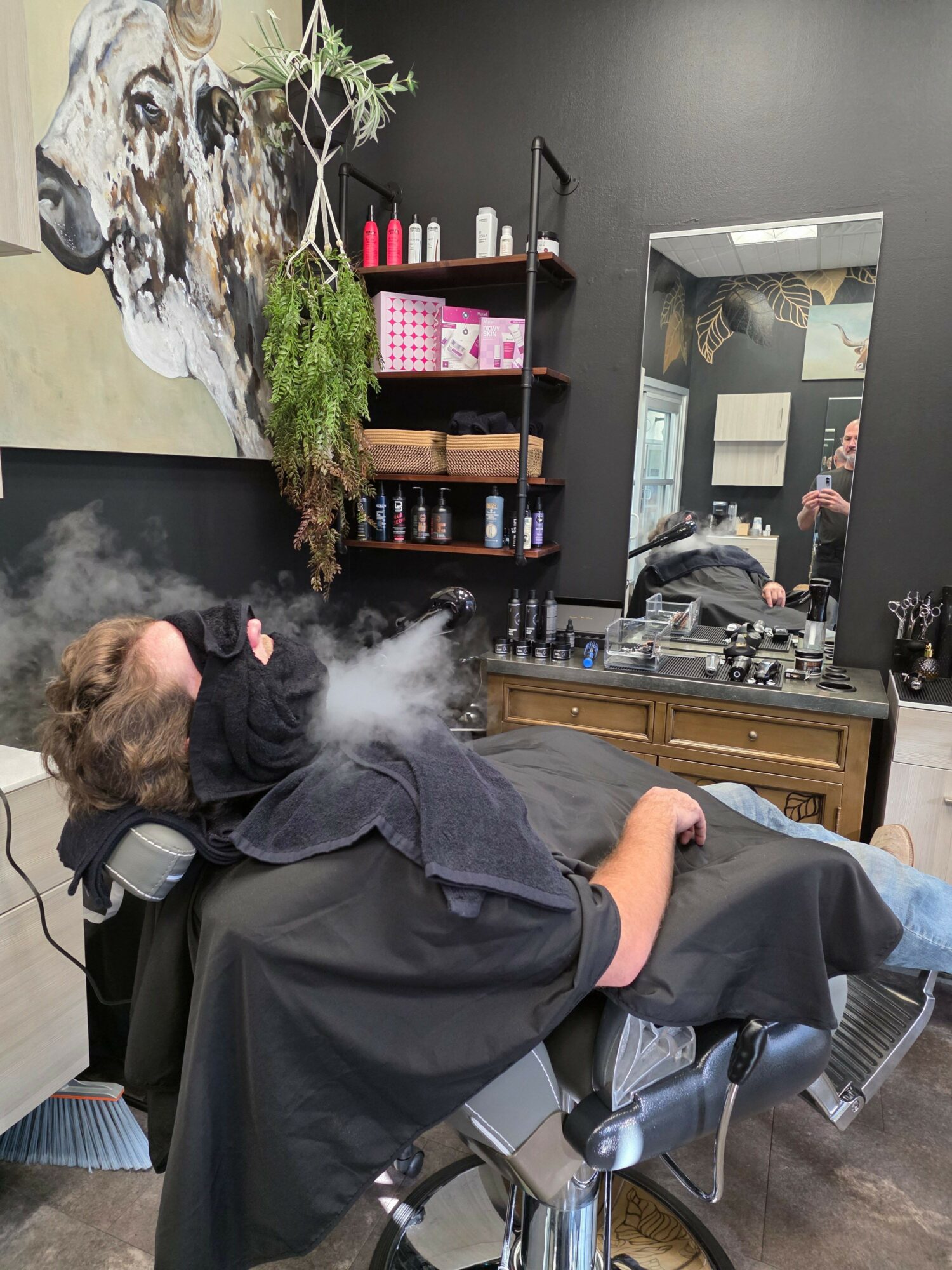 Person sitting in a salon chair with a black cape, getting a haircut, with a mirror and salon supplies in the background.
