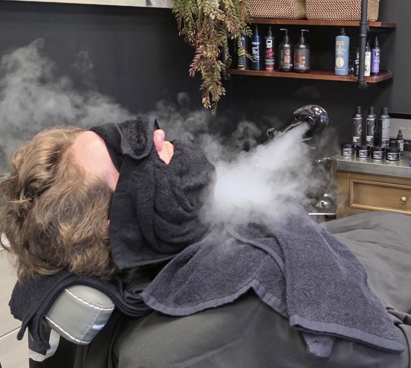 Person lying on a chair, vaping with vapor visible, in a room with shelves of bottles and plants, face covered with a cloth.