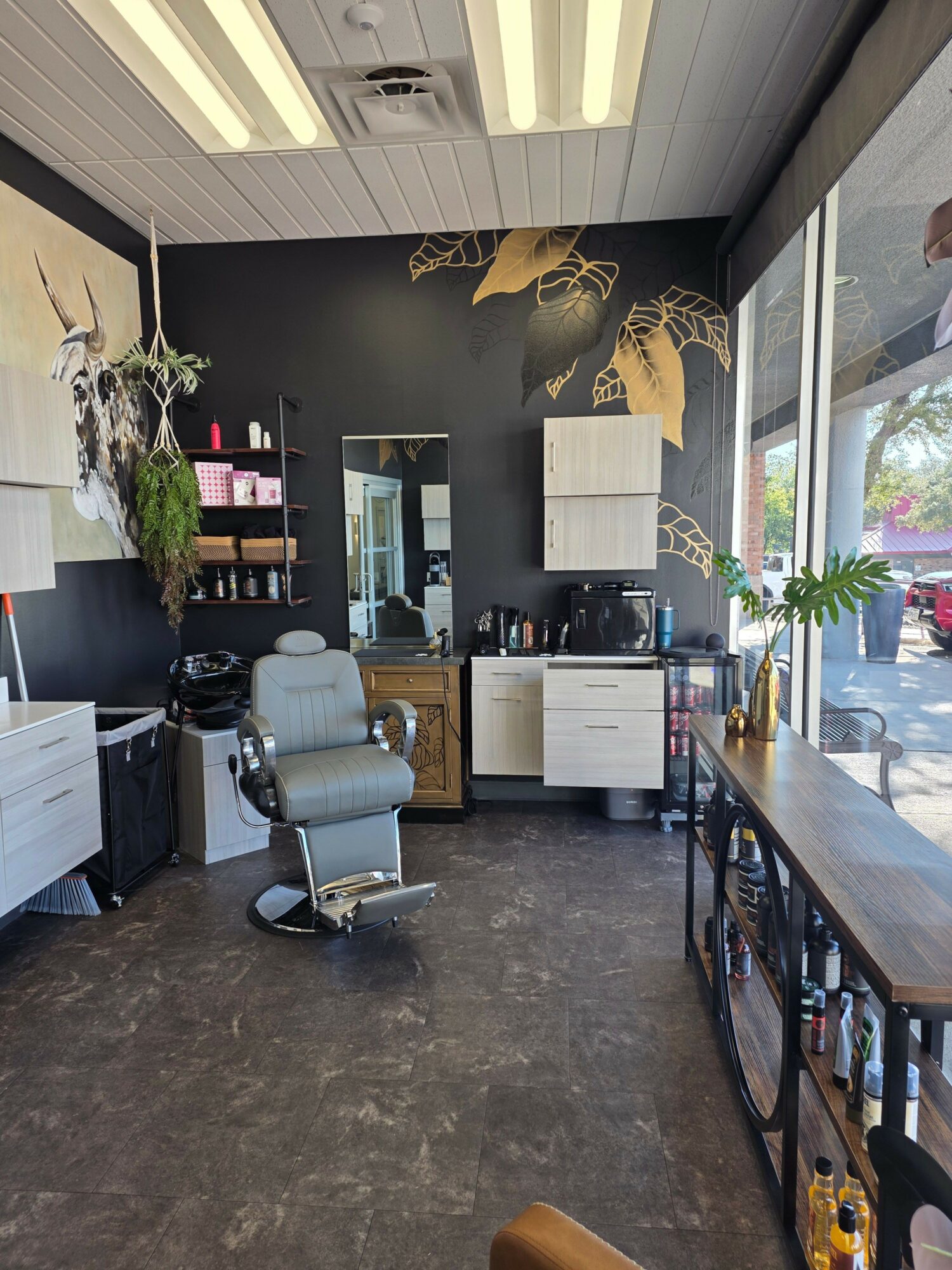 Salon with a styling chair, large window, plants, and shelves with hair products, black wall, and gray floor.