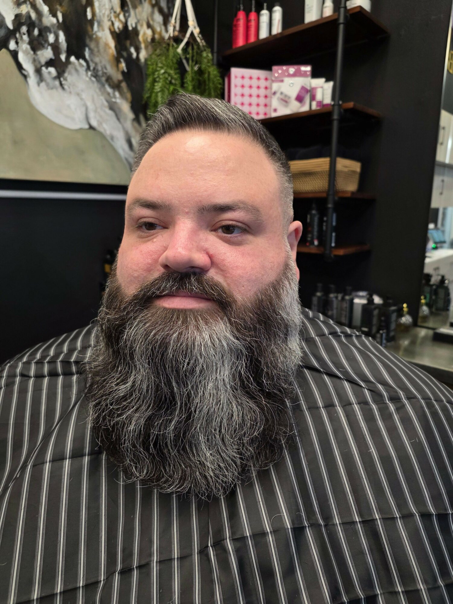Man with a long beard and mustache, sitting in a barber shop, wearing a striped cape, with shelves and plants in background.