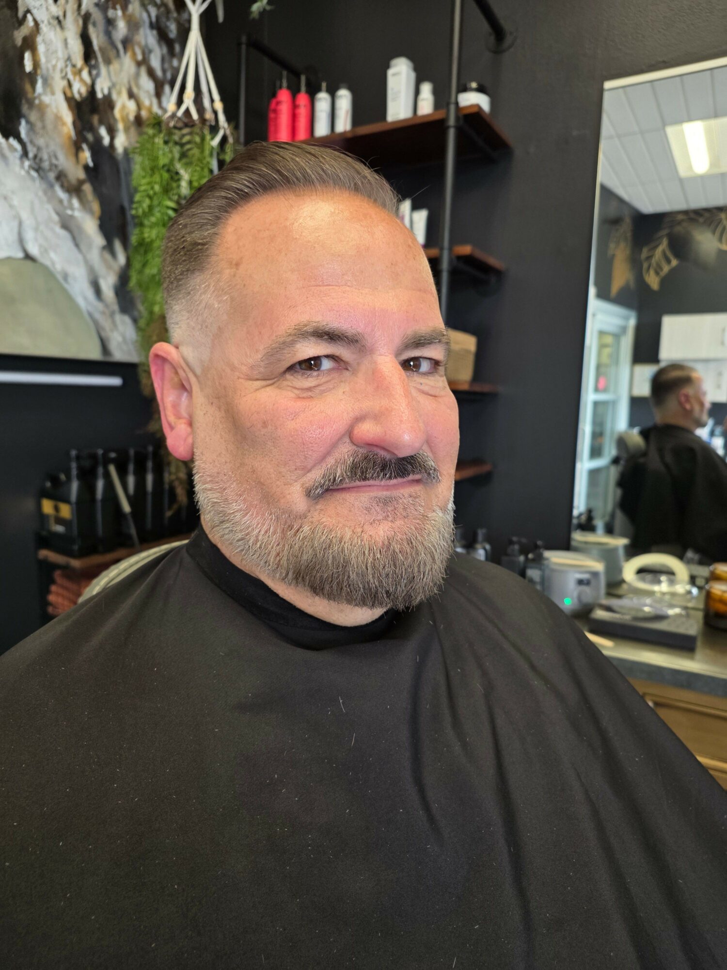 Man with short hair and beard sitting in a barbershop chair, looking at the camera.