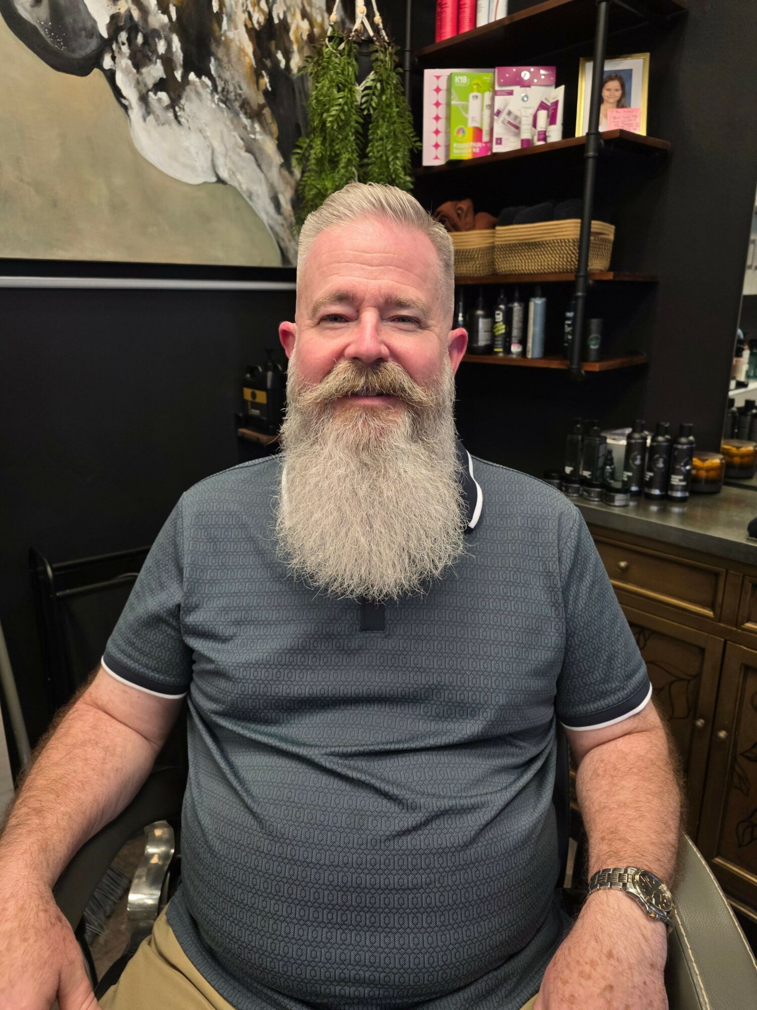 Man with a long white beard and short hair, wearing a gray shirt, sitting indoors with a black background and shelves.