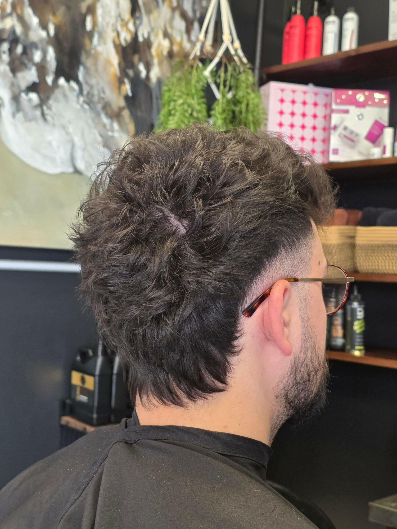Side view of a person with curly hair, wearing a black shirt, in a room with shelves and plants.